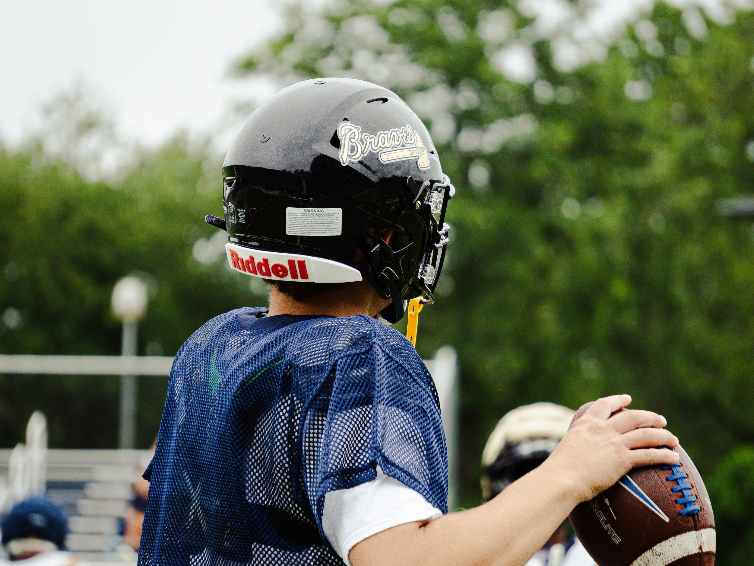 A young athlete wearing a football helmet with a Braves logo, holding a football, during an outdoor sports event with trees in the background.