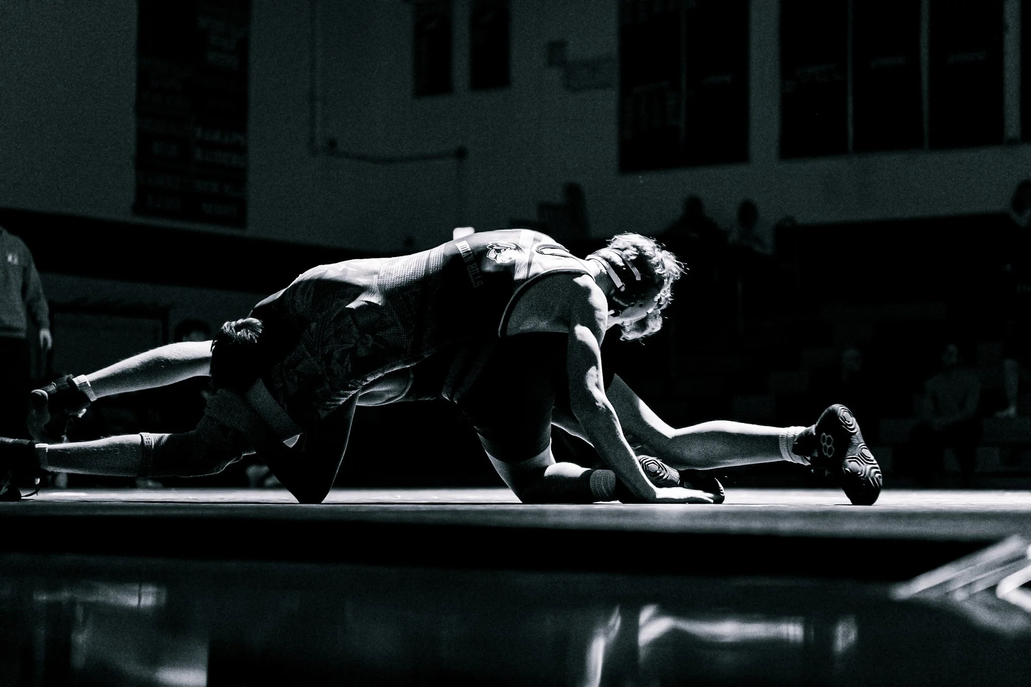 A black and white photo of wrestlers on the mat during a match, with a coach or referee overseeing in the background.