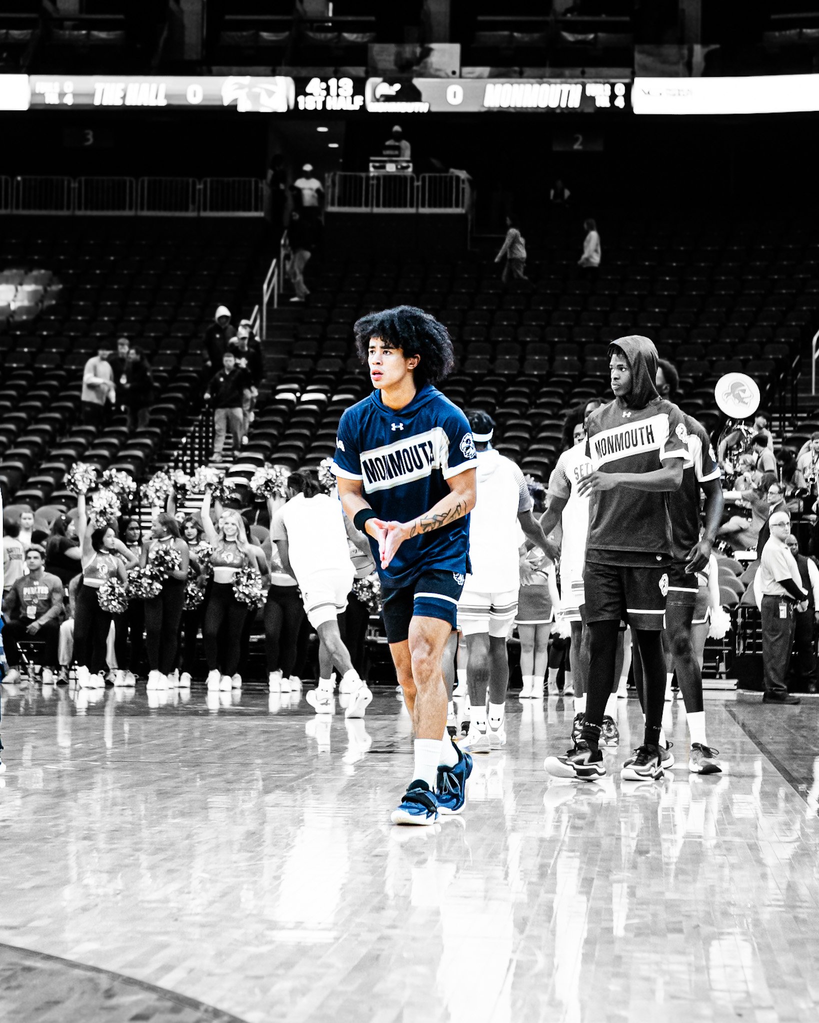 Basketball players warming up on the court before a game in an arena with a scoreboard overhead showing 4:19 left in the first half. The players are wearing jerseys that say 'Monmouth' and have a Black and white filter applied, with one player in ful