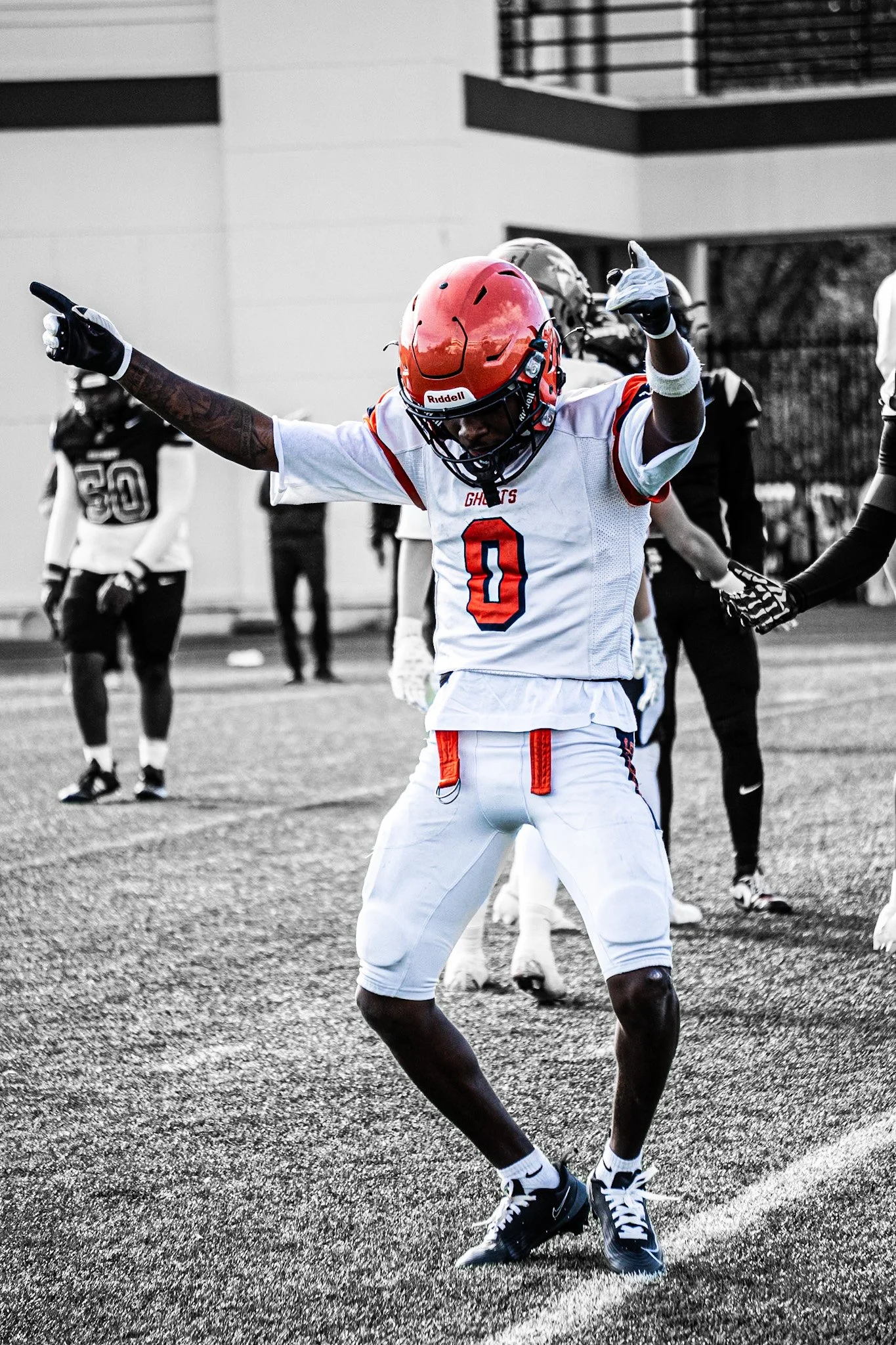 An American football player in a white uniform with red and blue accents, wearing a red helmet with a faceguard, celebrating on a football field with arms raised and pointing, others in black and white uniforms in the background.