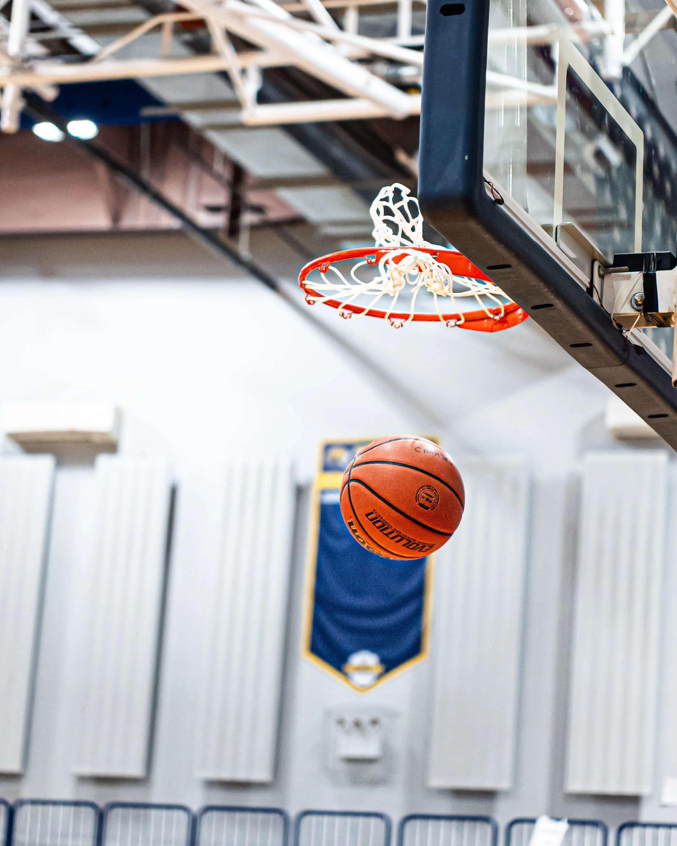A basketball hoop with a basketball passing underneath in an indoor gym.