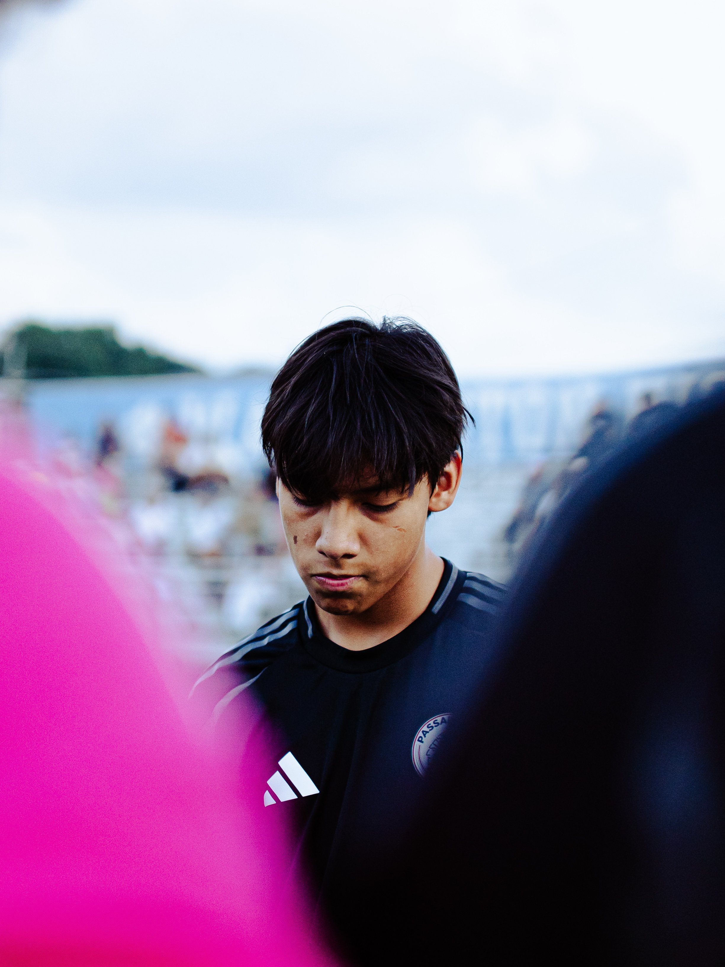 Close-up of a young man with dark hair and a football jersey, looking down, with a blurred outdoor background and pink crop on the left side.