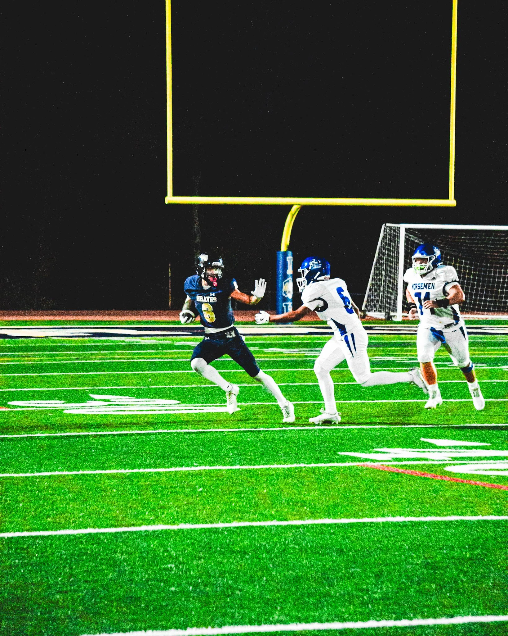 Nighttime American football game with players on the field, one player in a dark uniform appears to be catching the ball while another player in white tries to block or defend. A goalpost is visible in the background.