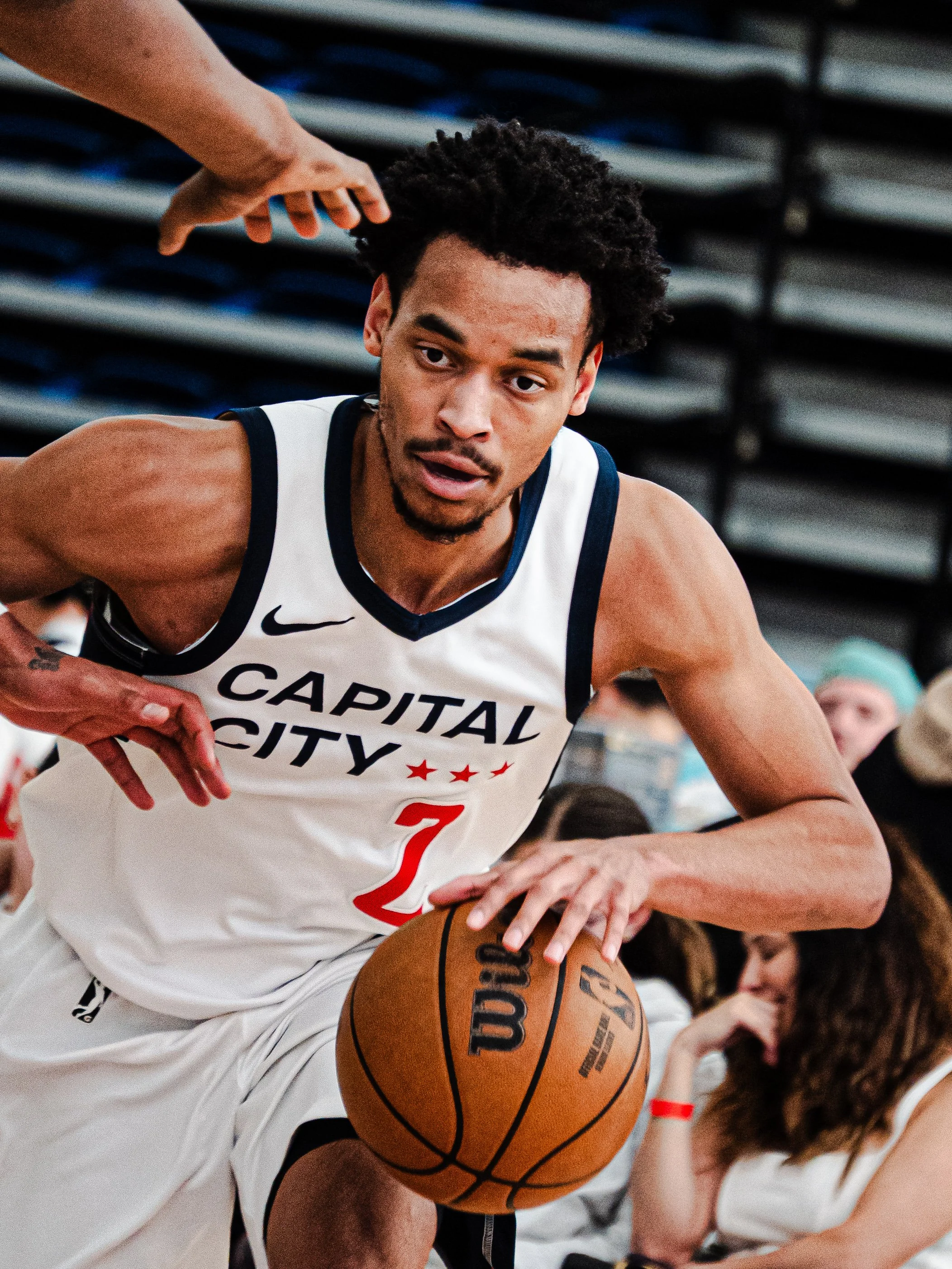 A male basketball player in a white and navy uniform with 'Capital City' written on it, wearing the number 7, is in the act of dribbling an orange Wilson basketball during a game. Someone's hand is reaching toward his head from above, possibly a refe