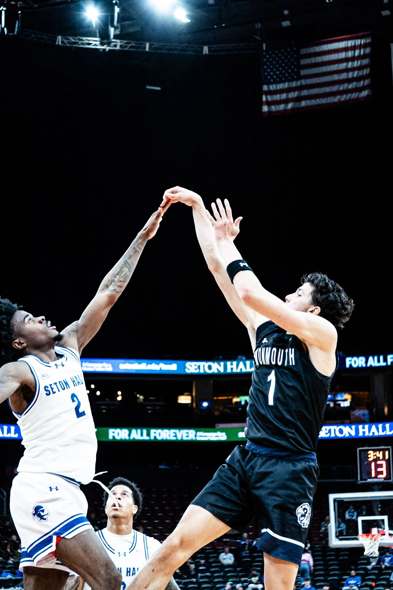 Two basketball players from opposing teams jumping to catch the ball in a game at Seton Hall, with one player wearing a white jersey and the other in black, under bright stadium lights and an American flag hanging in the background.