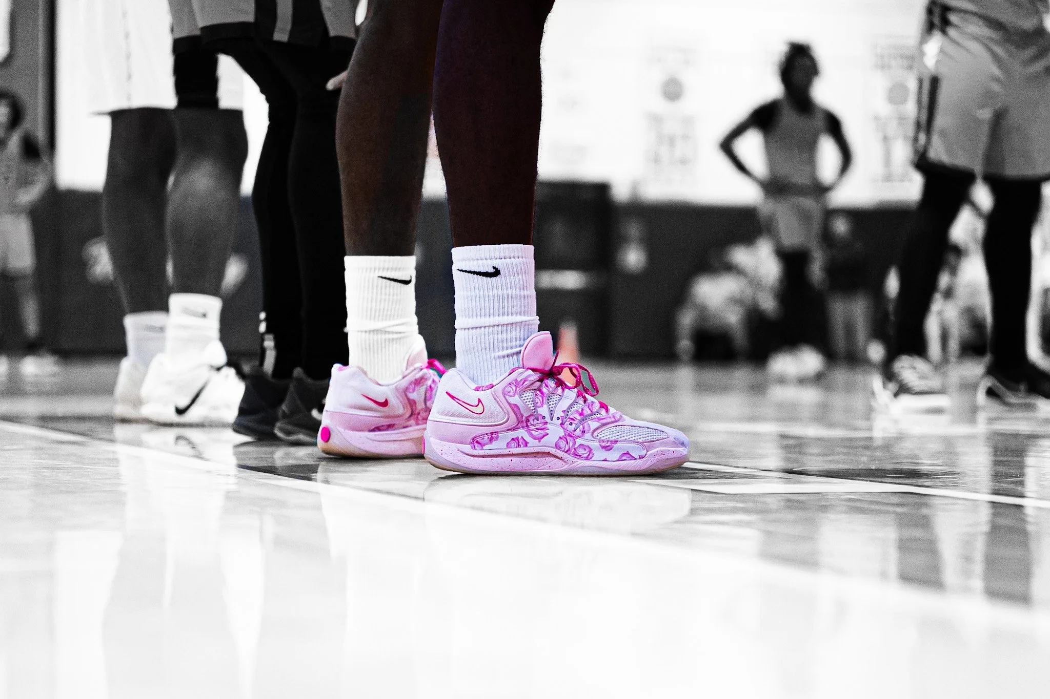 Close-up of a person wearing pink and white Nike athletic shoes and white socks with a Nike logo, standing on a gymnasium floor during a sports game. Other players and spectators are visible in the blurred background.