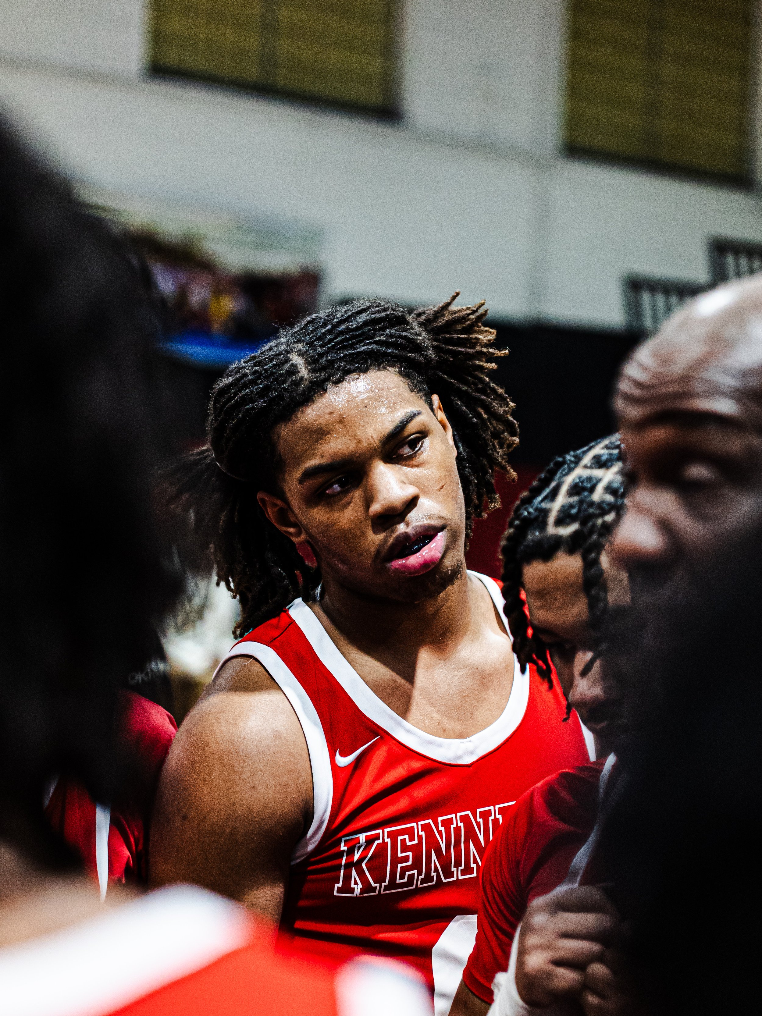 A female basketball player in a red and white jersey with the word "Kentucky" is in a huddle with teammates during a game or team meeting.