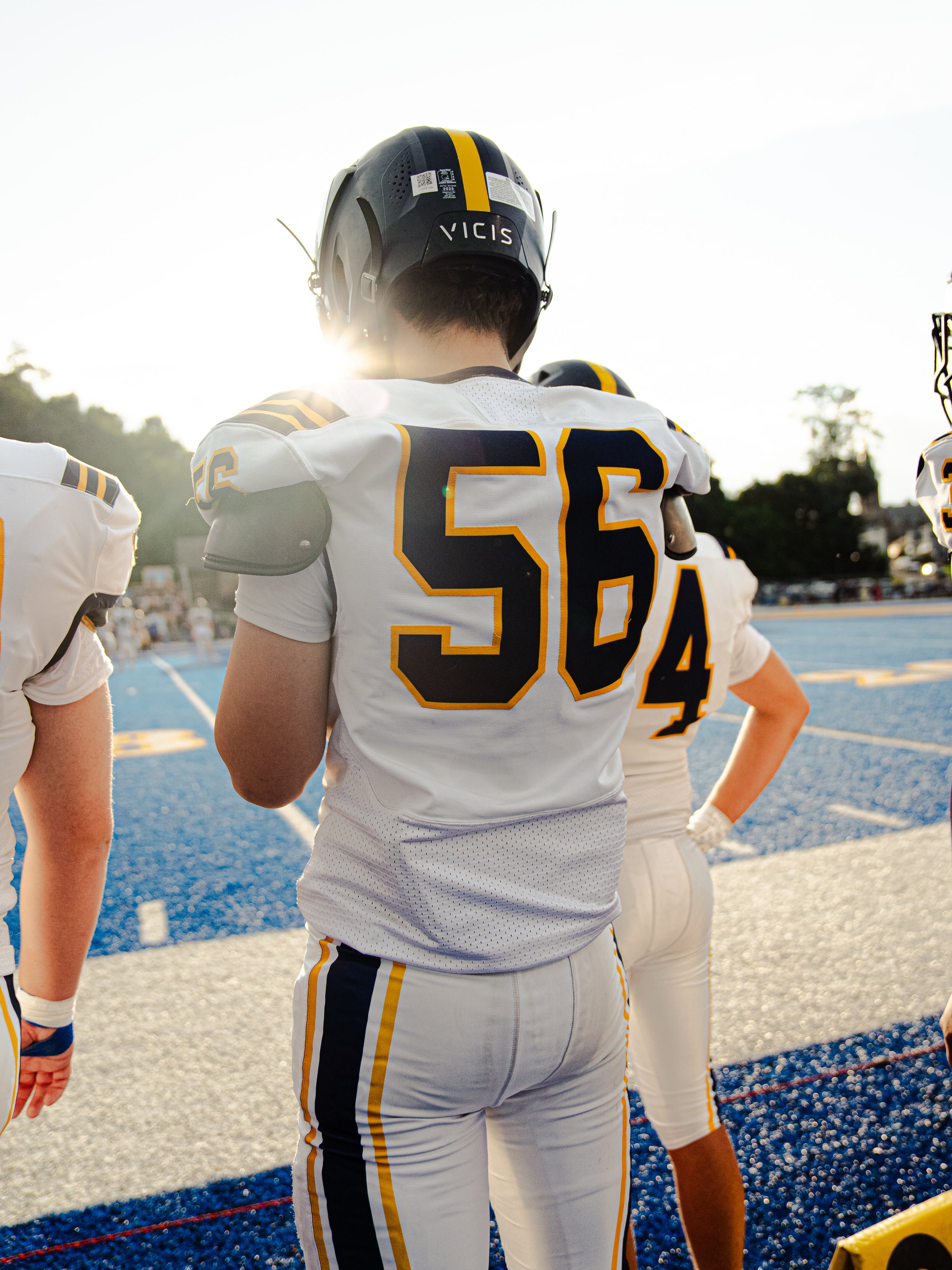 Football players in white uniforms with black numbers, standing on a bright blue field with a sideline marked by a red rope, during sunset or late afternoon.
