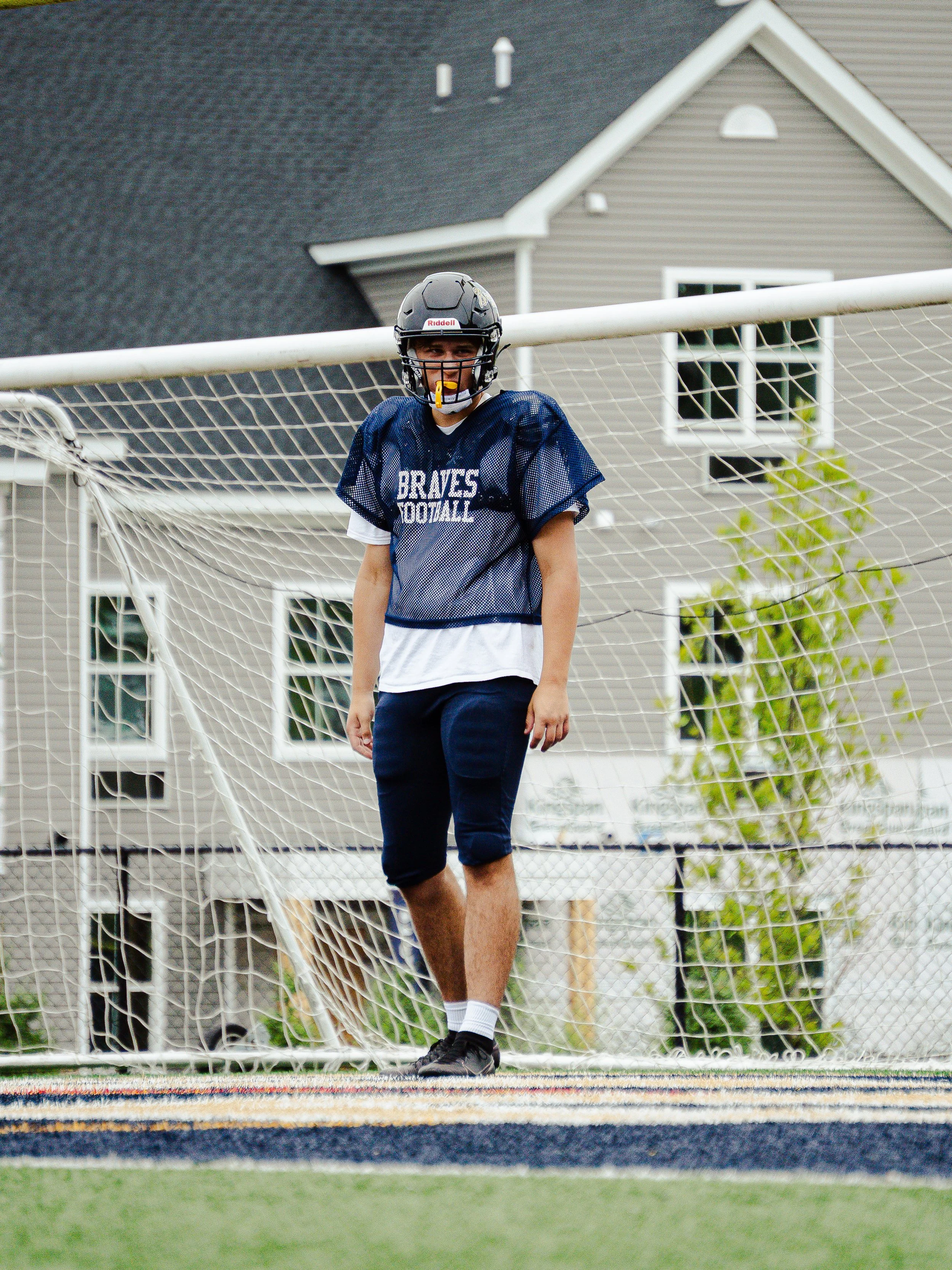 A young football player standing on a field behind a goal net, wearing a helmet, blue practice jersey, and athletic shorts, with houses visible in the background.