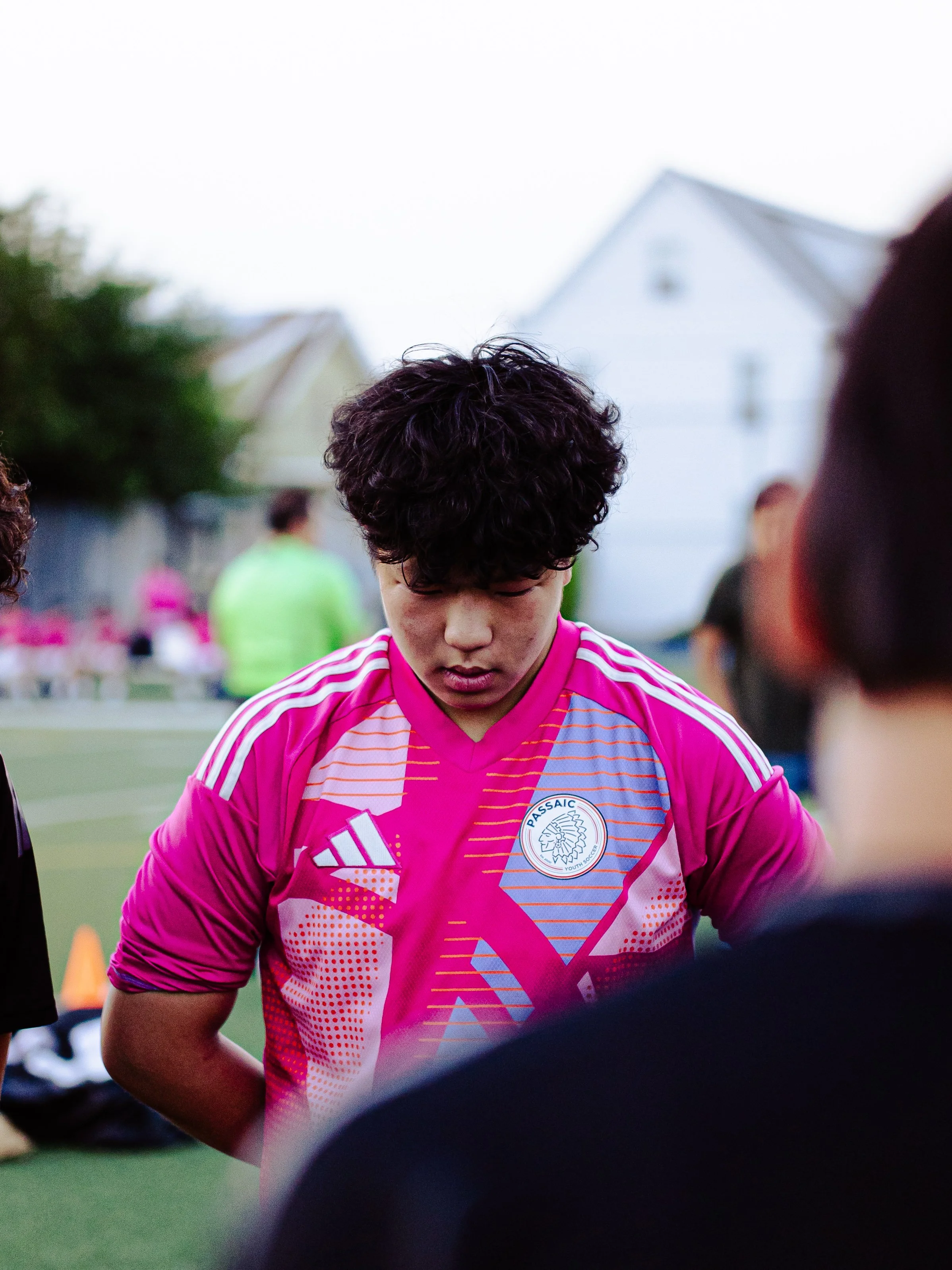 A young male soccer player wearing a pink and purple sports jersey with a logo that reads 'PASSAIC YOUTH SOCCER' on his chest, standing outdoors on a sports field with houses in the background.