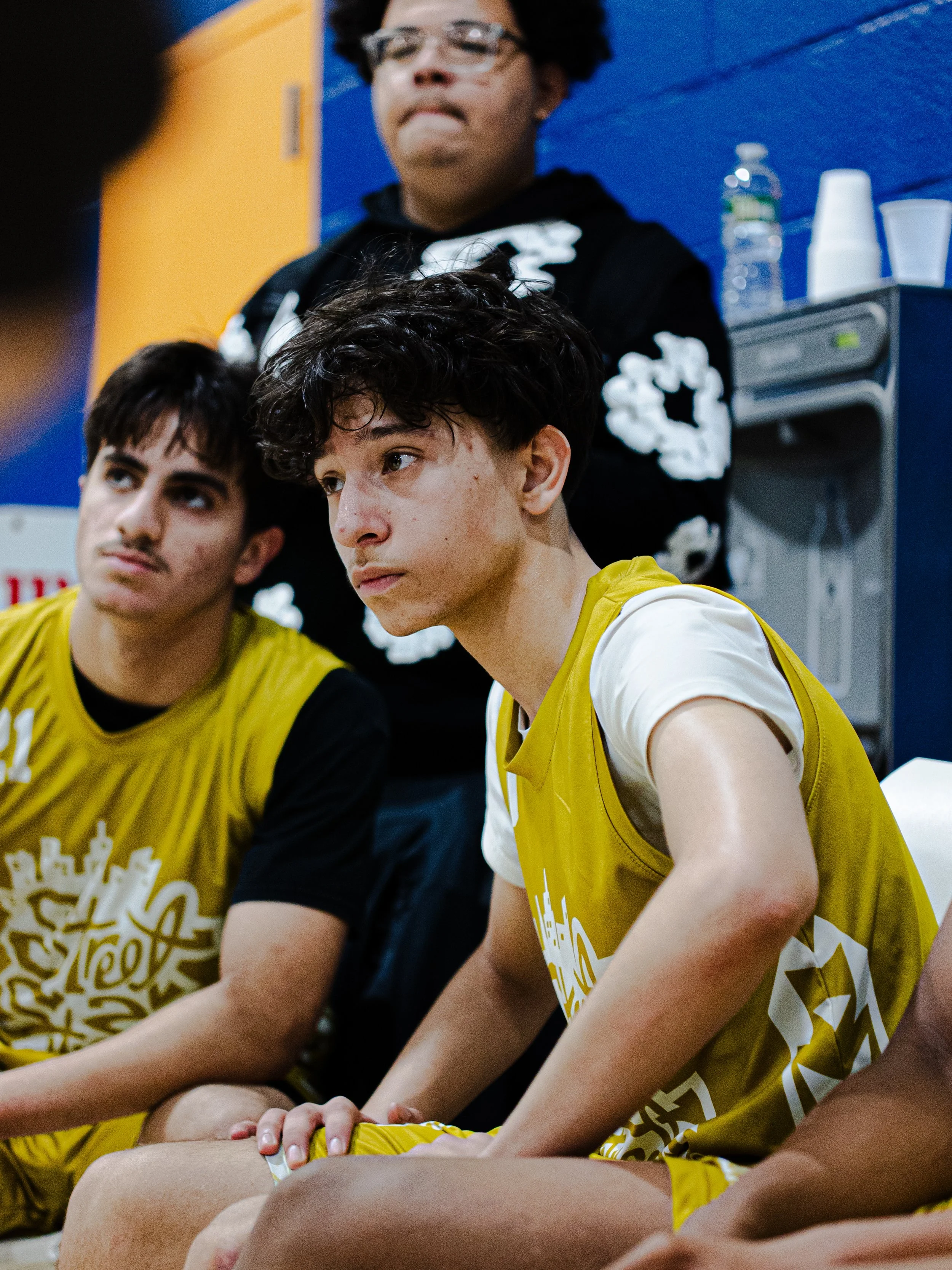 Group of young men, wearing yellow sports jerseys, sitting on the floor indoors. One young man in the foreground has curly dark hair, fair skin, and is looking to the right. Behind him, a young man with straight dark hair and a darker skin tone is lo