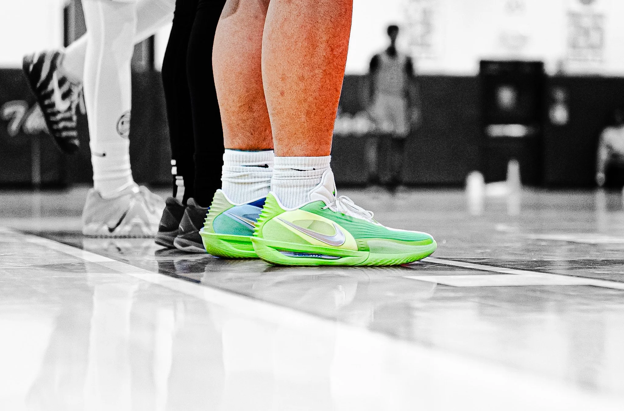 Close-up of a person wearing neon green Nike sneakers and white socks, standing on a basketball court with other players visible in the background.
