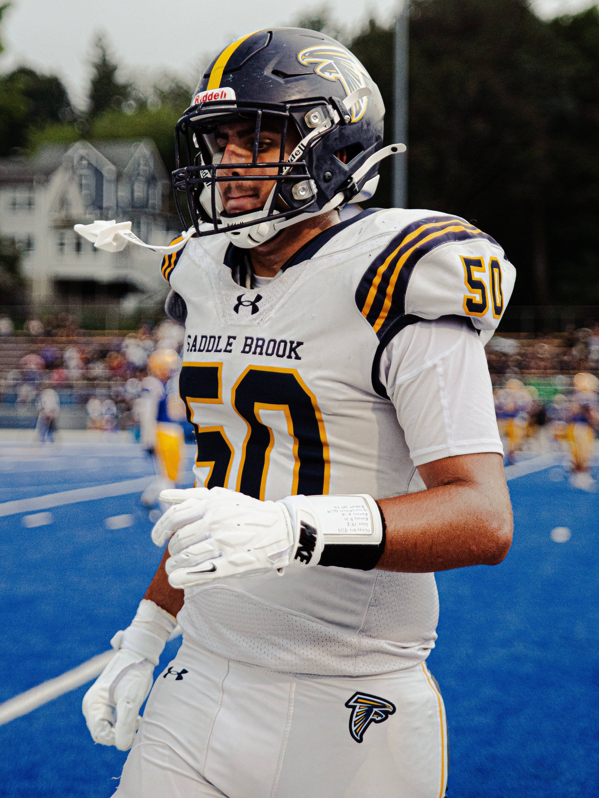 A football player from the Saddle Brook team wearing a white jersey, black and yellow helmet, and white gloves standing on a football field with a blue surface during a game or practice.