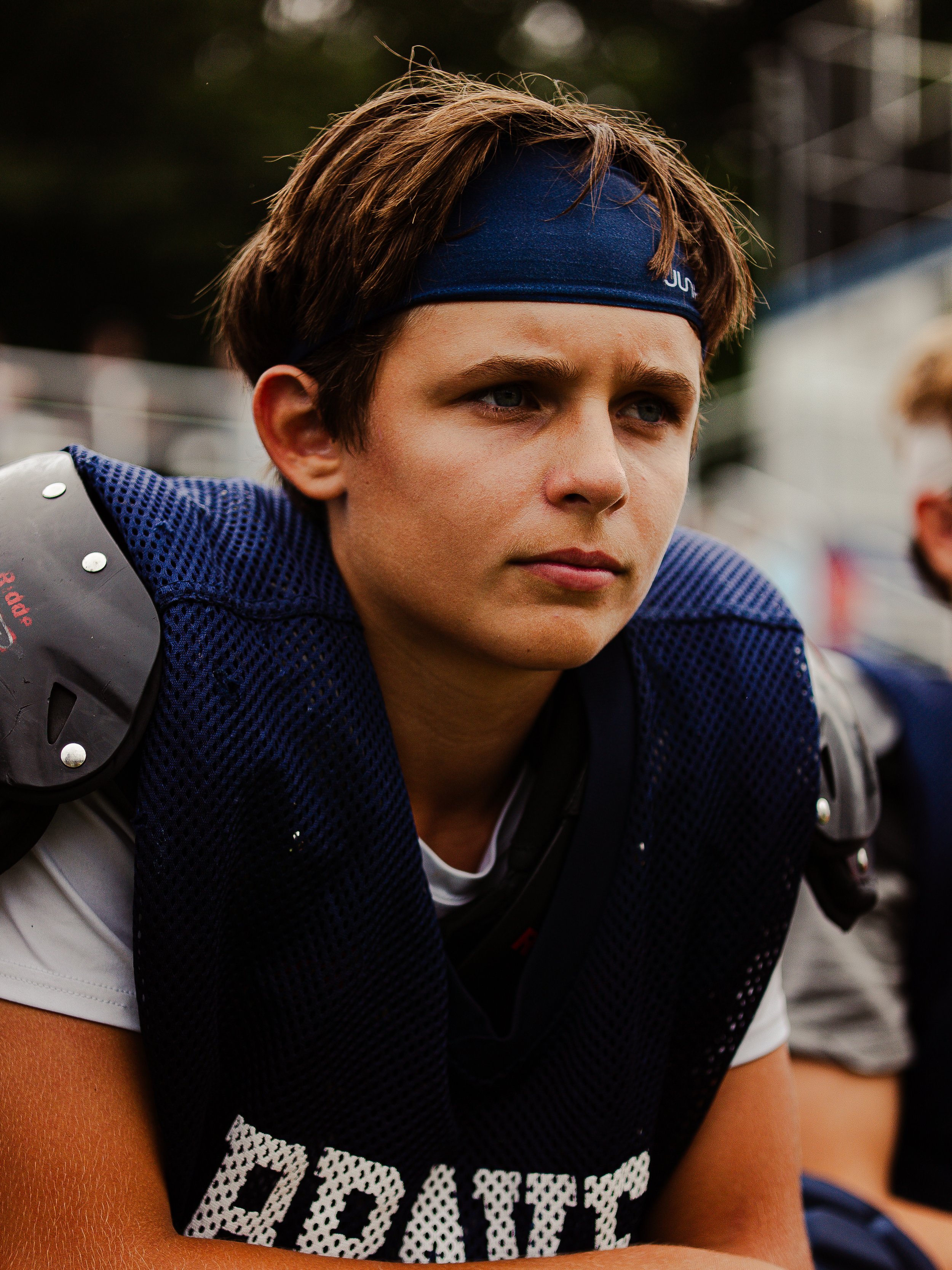 Close-up of a young person dressed in football gear, including a dark blue headband, a black and white jersey with the word 'DOMINATE' on the front, and shoulder pads, sitting outdoors with a focused expression.