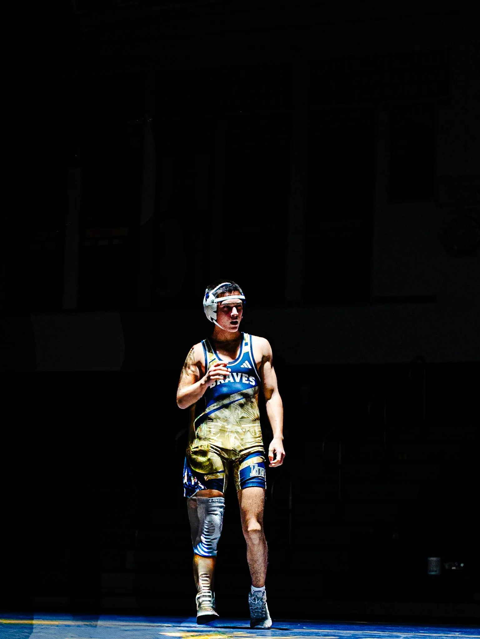 A male wrestler in a blue and yellow singlet walking on a wrestling mat, wearing a protective headgear and sports shoe, with a dark background and the spotlight on him.