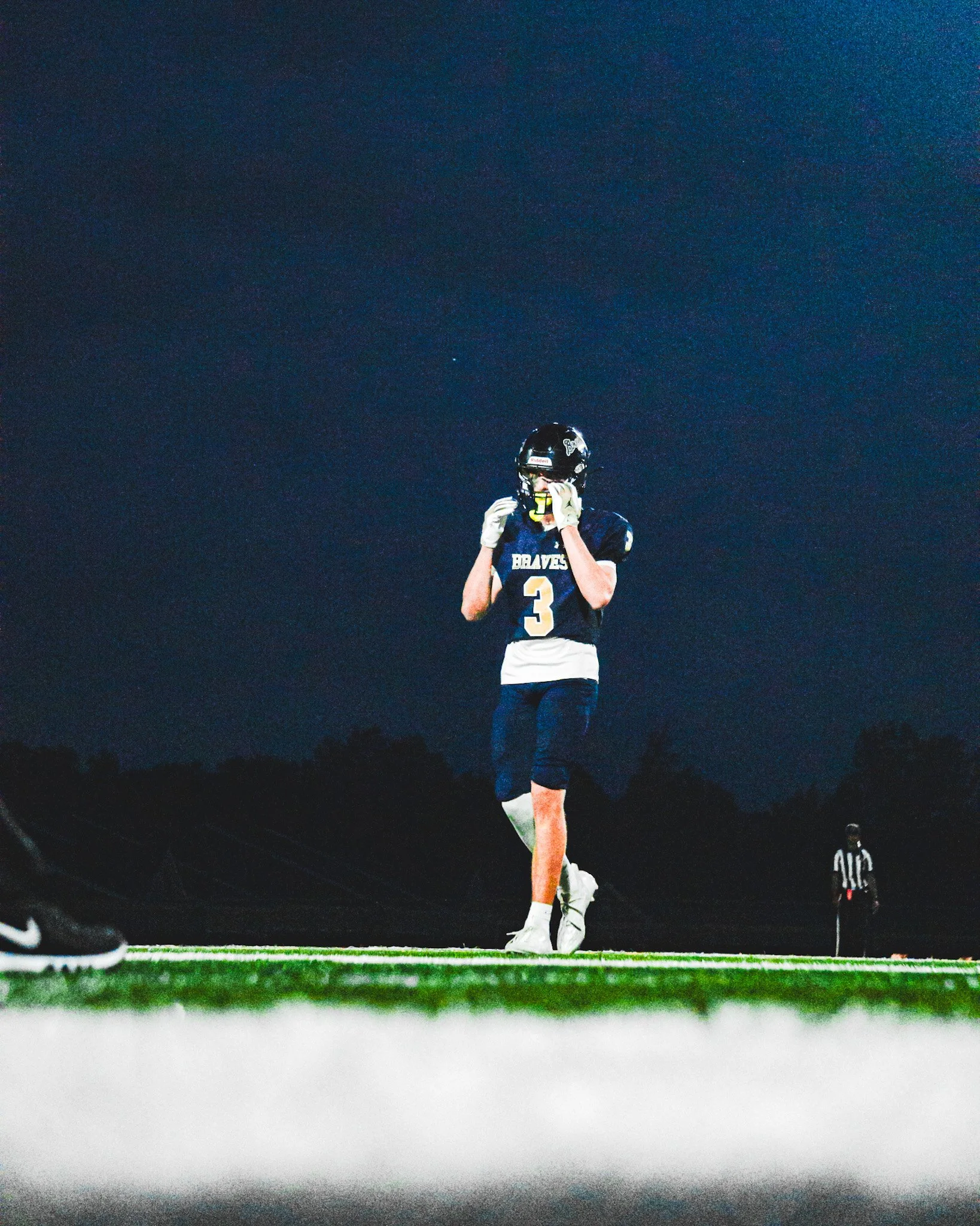 A football player wearing a black helmet and navy jersey with the number 3 is standing on the field under a dark sky, adjusting his helmet. Another player dressed similarly is partially visible on the side and a referee is in the background.