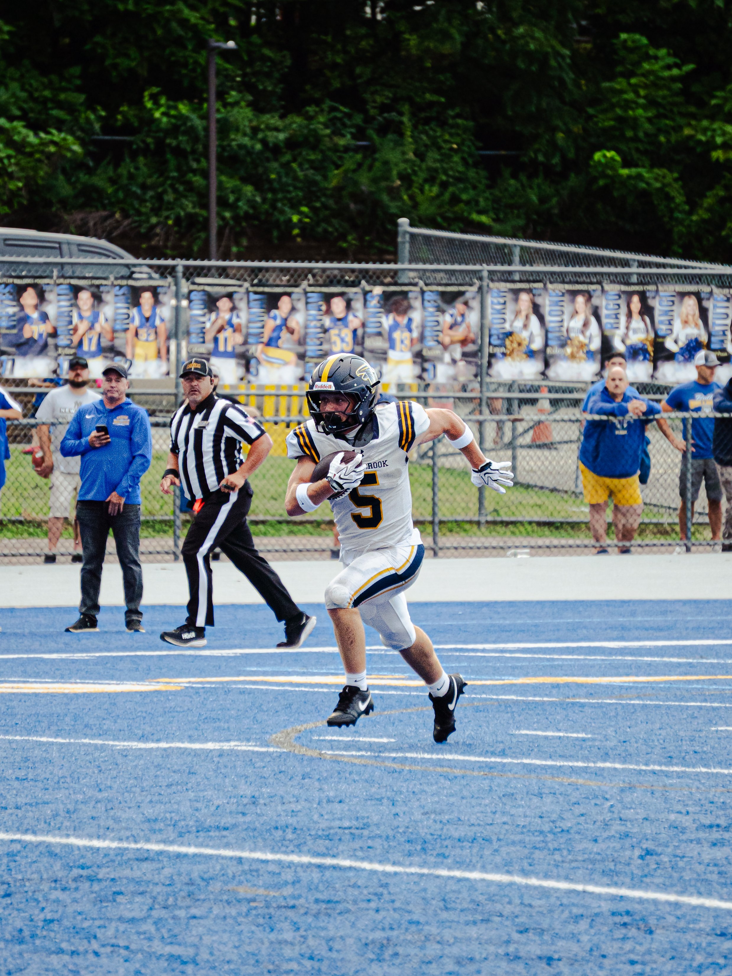 A football player wearing a helmet and jersey with the number 5 running on a blue track during a game. There are officials, coaches, and spectators behind a fence, with some holding phones and cheerleading banners in the background.