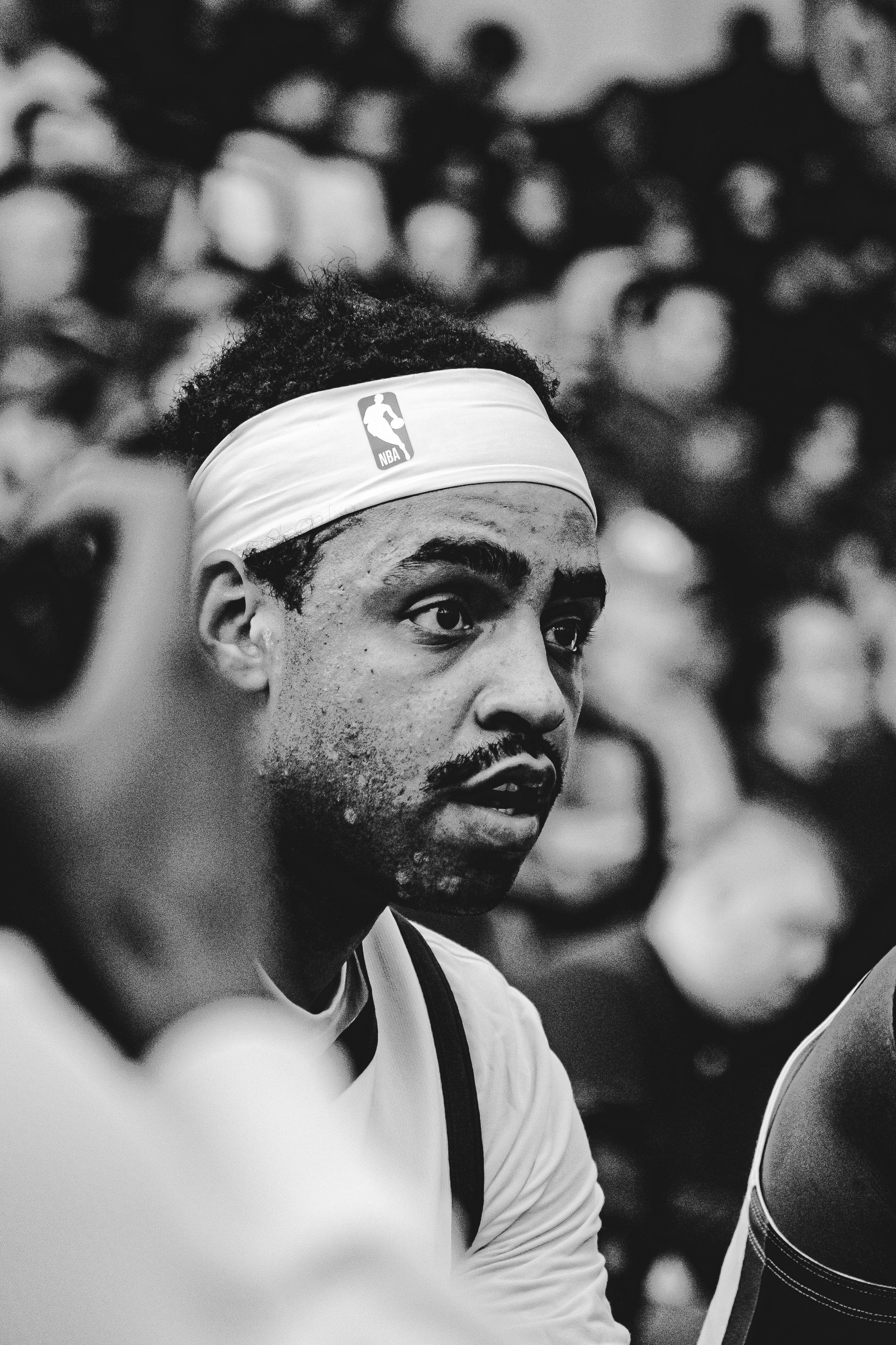 A black and white close-up of a male basketball player wearing a NBA headband, with an intense expression, sitting among other players.