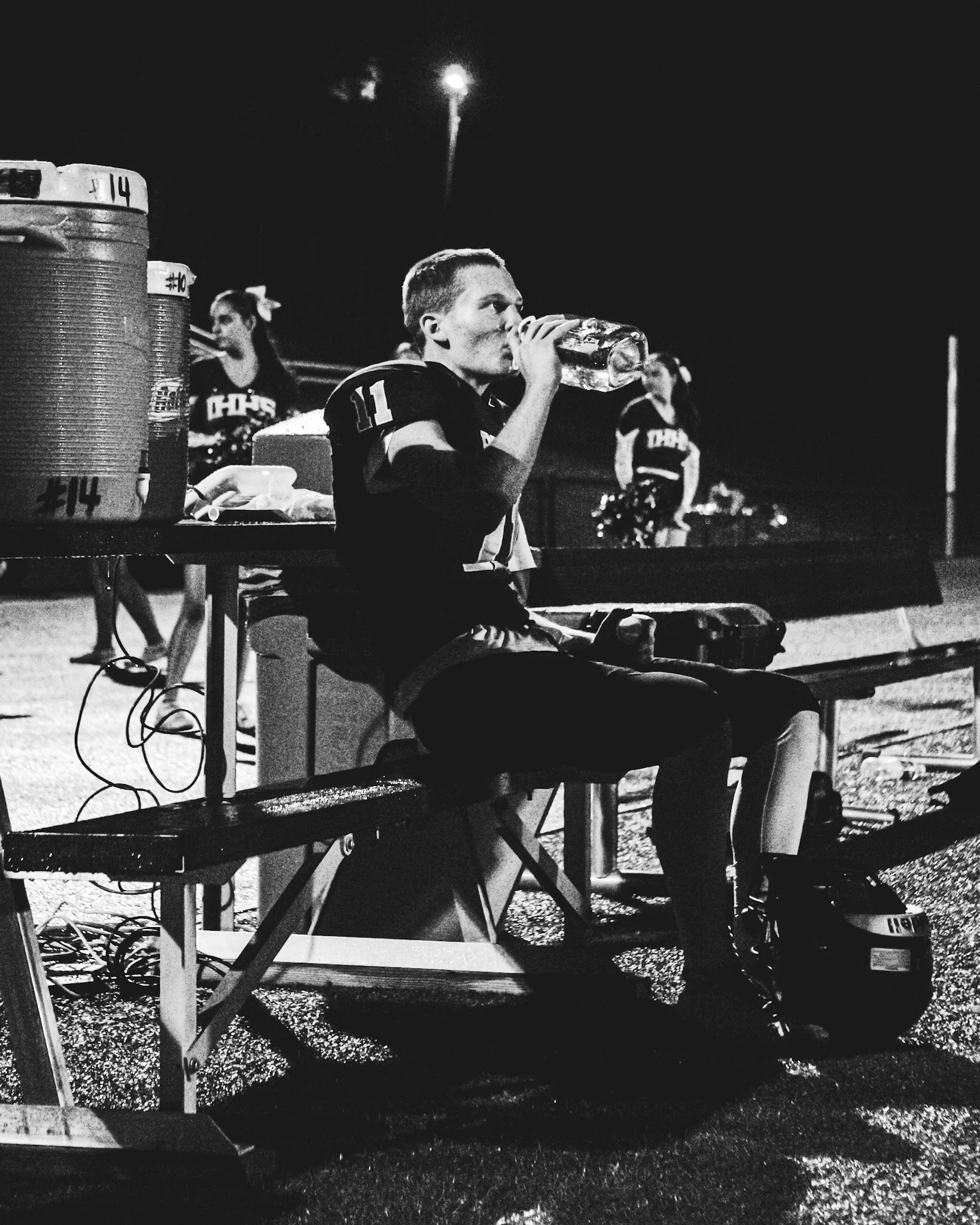 A night scene featuring a young man in a football uniform sitting on a bench, drinking from a large bottle, with other players and equipment in the background, illuminated by bright stadium lights.