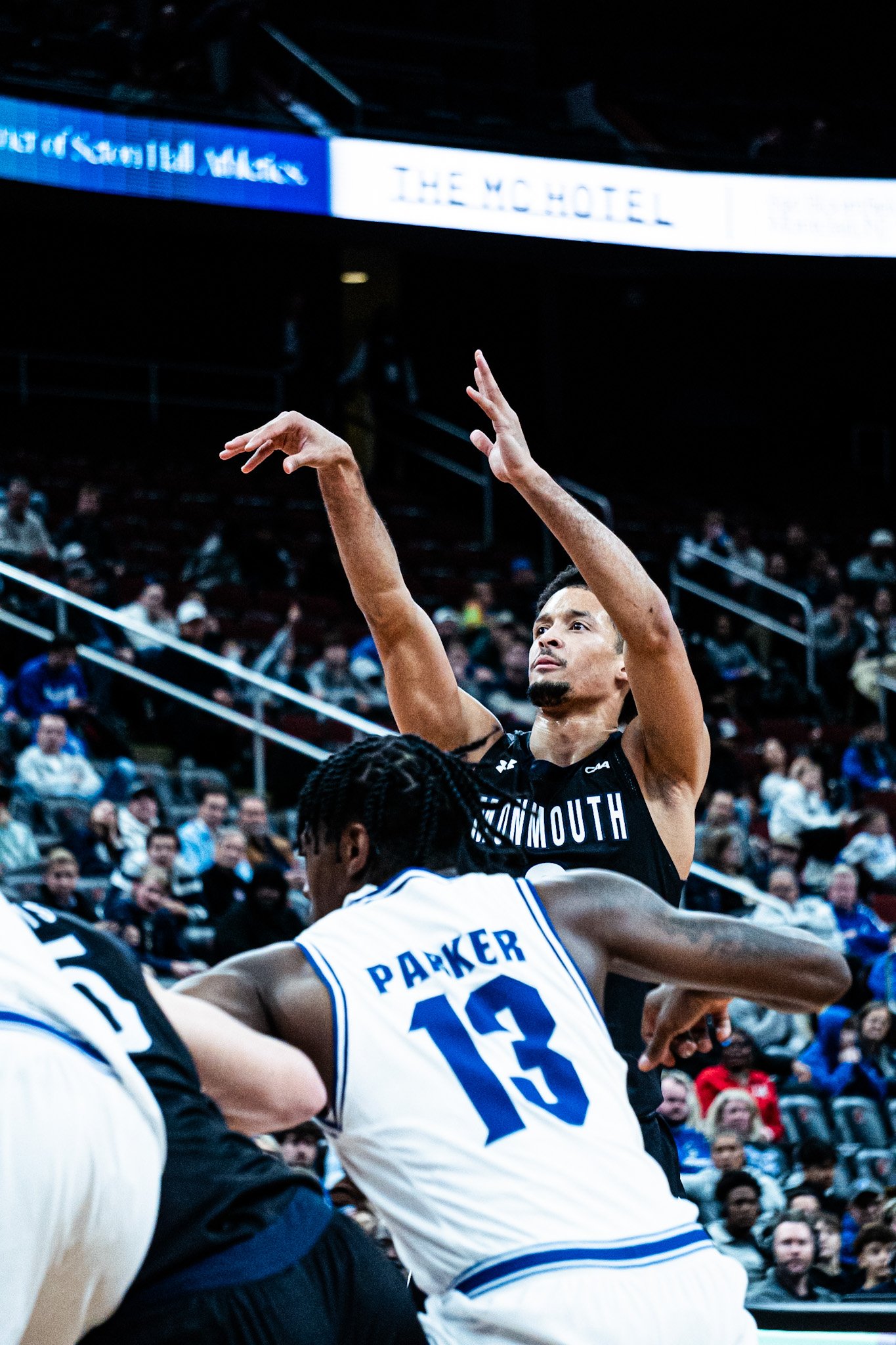 A basketball game with players in motion, including a player from Drexel University wearing a white jersey with blue accents and the name Parker, and a player from Monmouth University in a black jersey. The game is in an arena with an audience in the