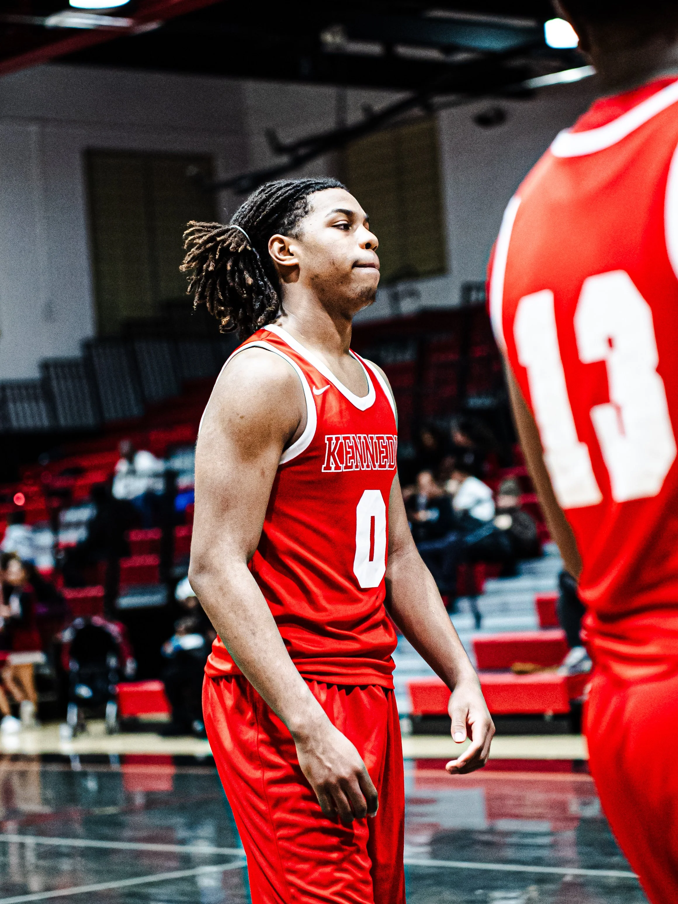 A basketball player in a red jersey with white trim and 'Kennedy' written on it is standing on a basketball court. The player has dreadlocks and a focused expression. Other players and spectators are visible in the background.