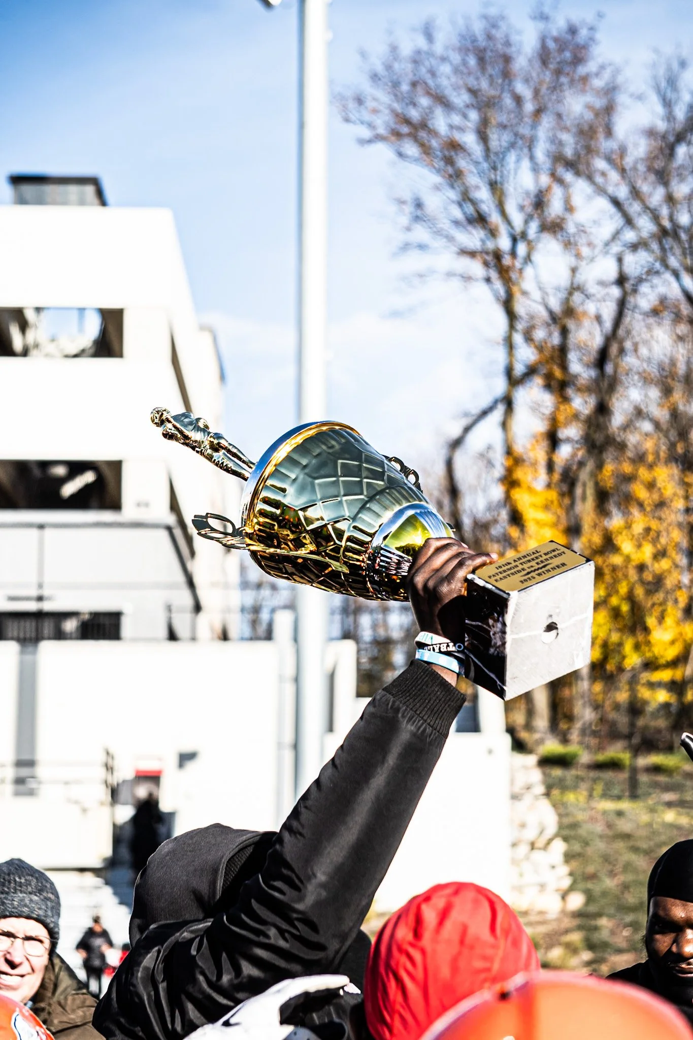 A person holding up a gold trophy during a daytime outdoor event surrounded by other people, some wearing warm clothing and beanies, with a building and trees in the background.