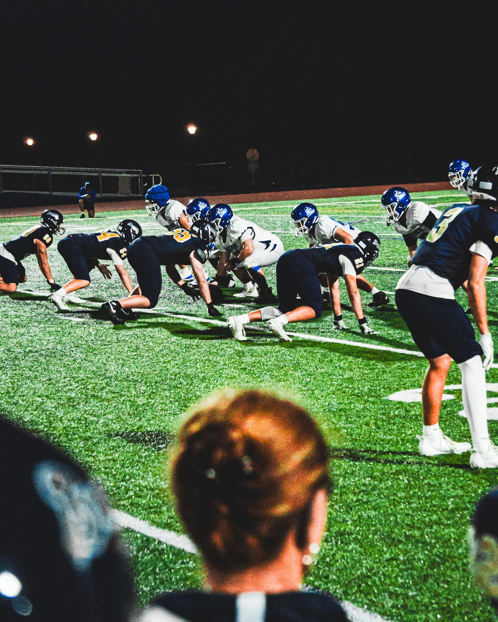 Nighttime photo of a youth football game with players on the field in formation, wearing helmets and uniforms. Bright lights illuminate the field, with a chain-link fence and empty stands in the background.