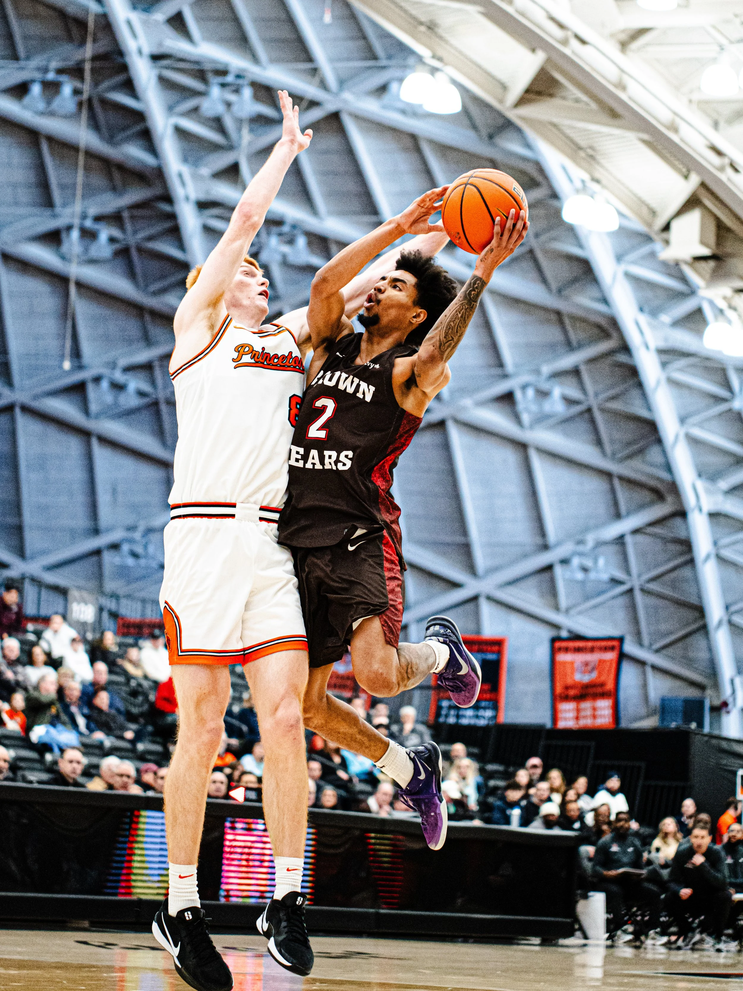 Two basketball players in mid-air during a game, with one attempting a shot while the other defends. The player in black is wearing jersey number 2, and the player in white is wearing jersey number 8. The scene takes place inside a sports arena with 
