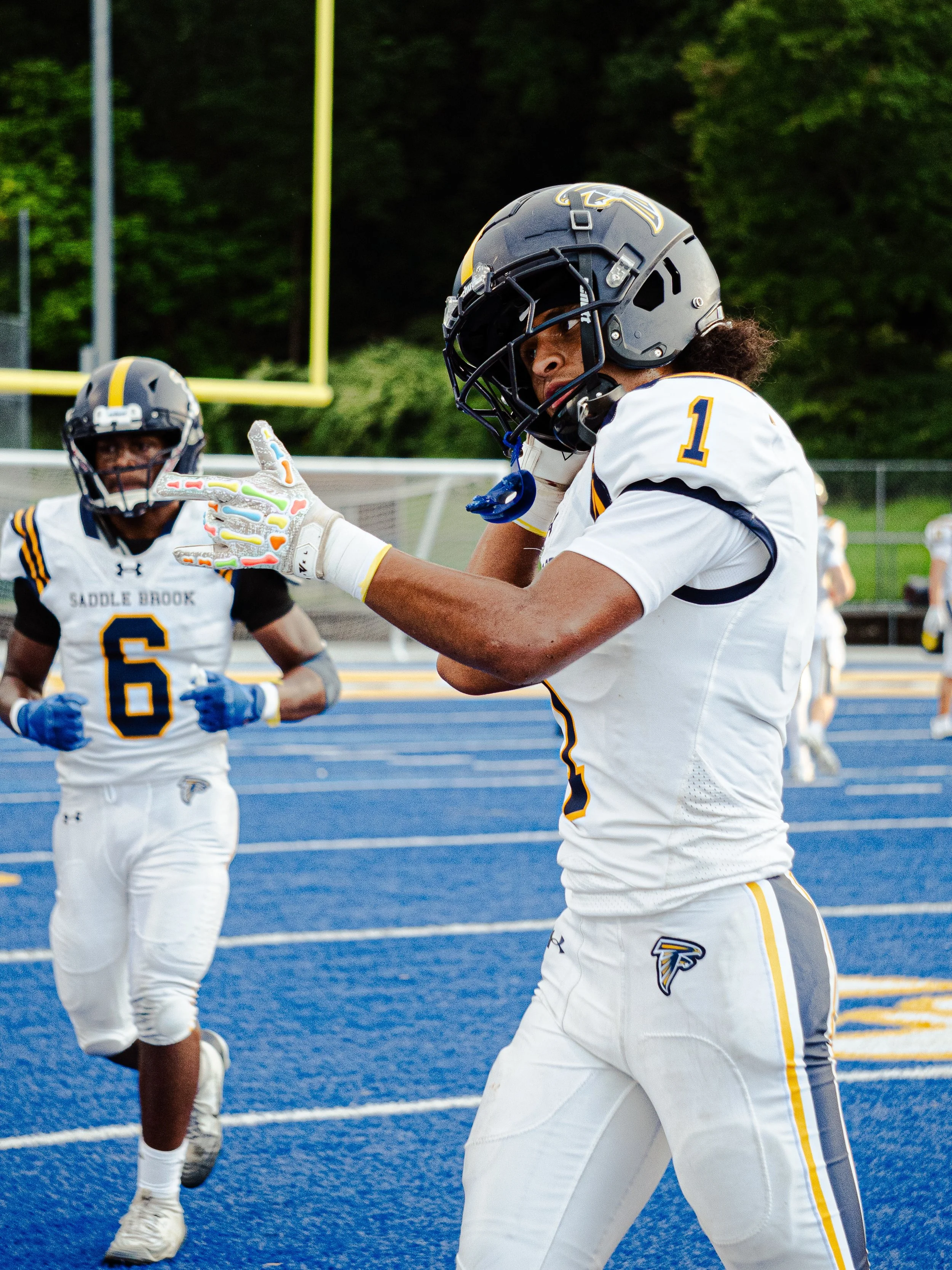 Two football players in white and navy uniforms with yellow accents, wearing helmets, standing on a blue running track with a yellow goalpost and green trees in the background.
