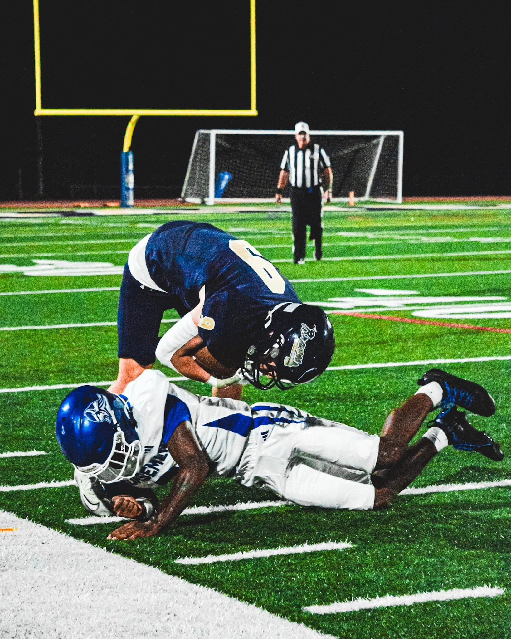 A football game at night with a player on the ground in a white and blue uniform, a player in a dark uniform kneeling nearby, a referee standing in the background, and a football goalpost on a brightly lit field.