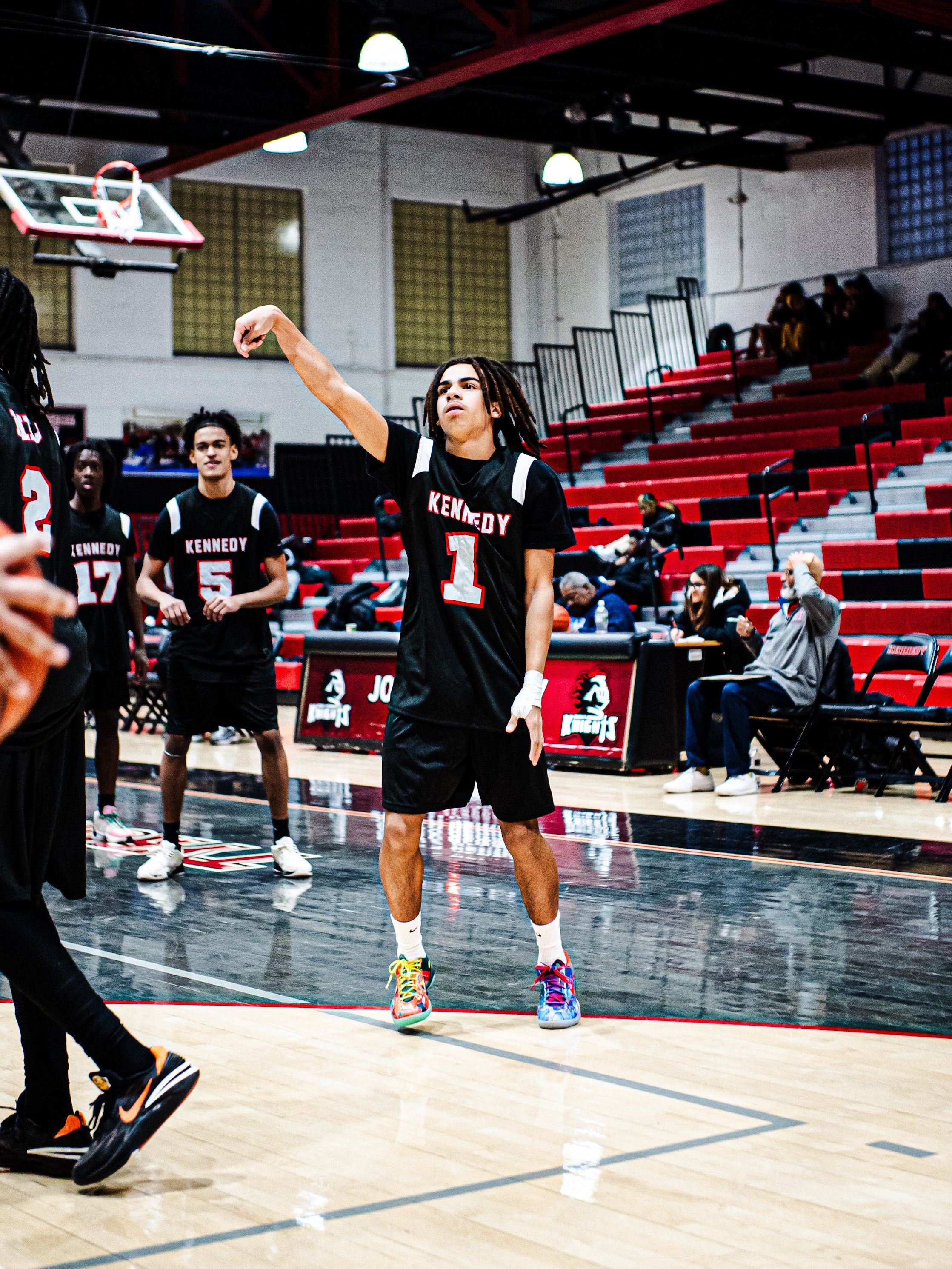 A young male basketball player with dreadlocks in a black jersey with red and white accents, labeled 'Kennedy 1', practicing free throws in an indoor gymnasium, with teammates and spectators watching.