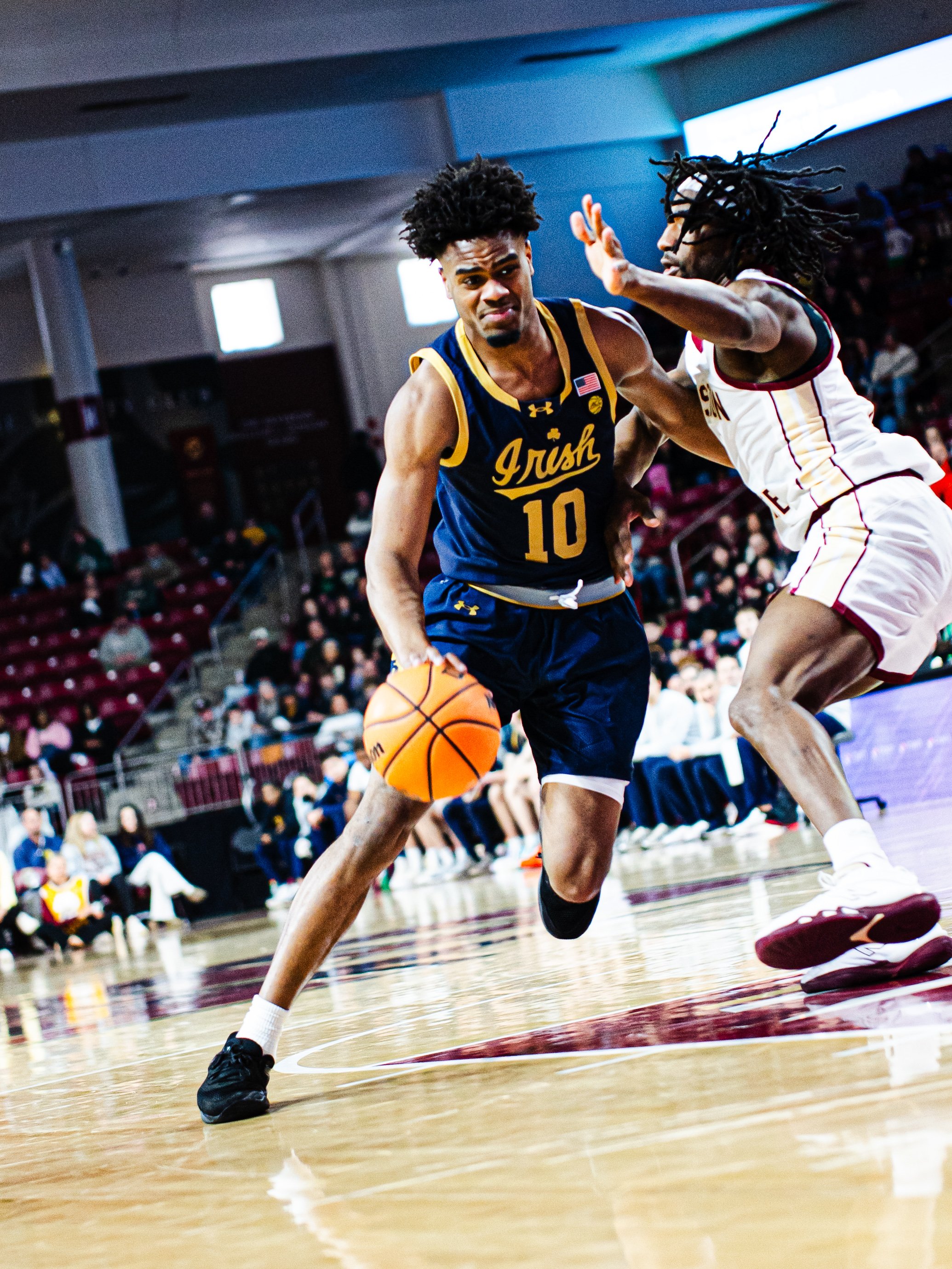Two basketball players are competing for the ball on the court during a game. The player in dark blue and yellow jersey with the number 10 is dribbling, while the other player in white with red and yellow accents is defending. The game takes place in