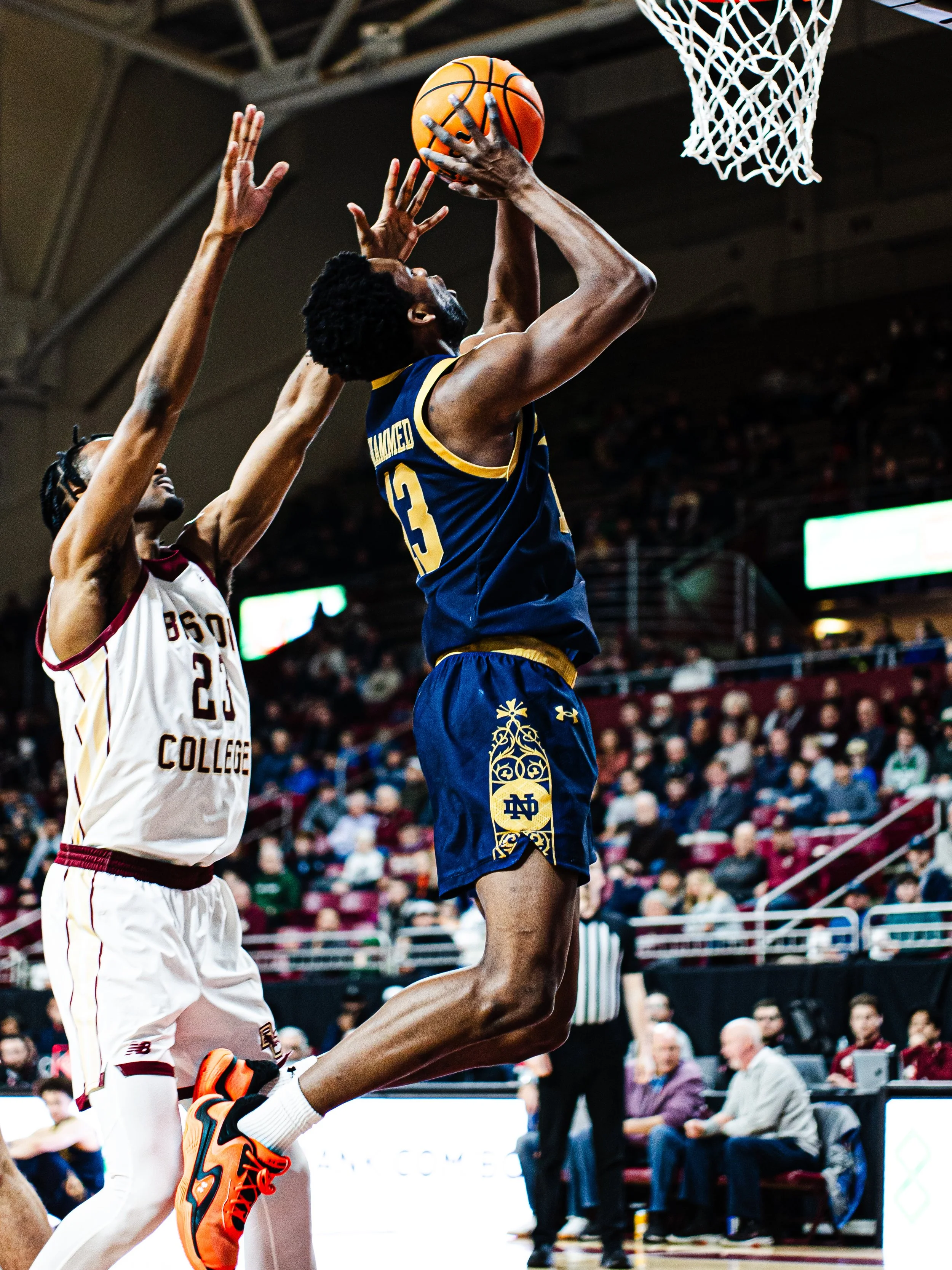 A basketball player from Notre Dame in blue and yellow uniform jumping to attempt a shot near the basket, while an opposing player in white and maroon jersey tries to block the shot. The scene takes place inside a crowded basketball arena.