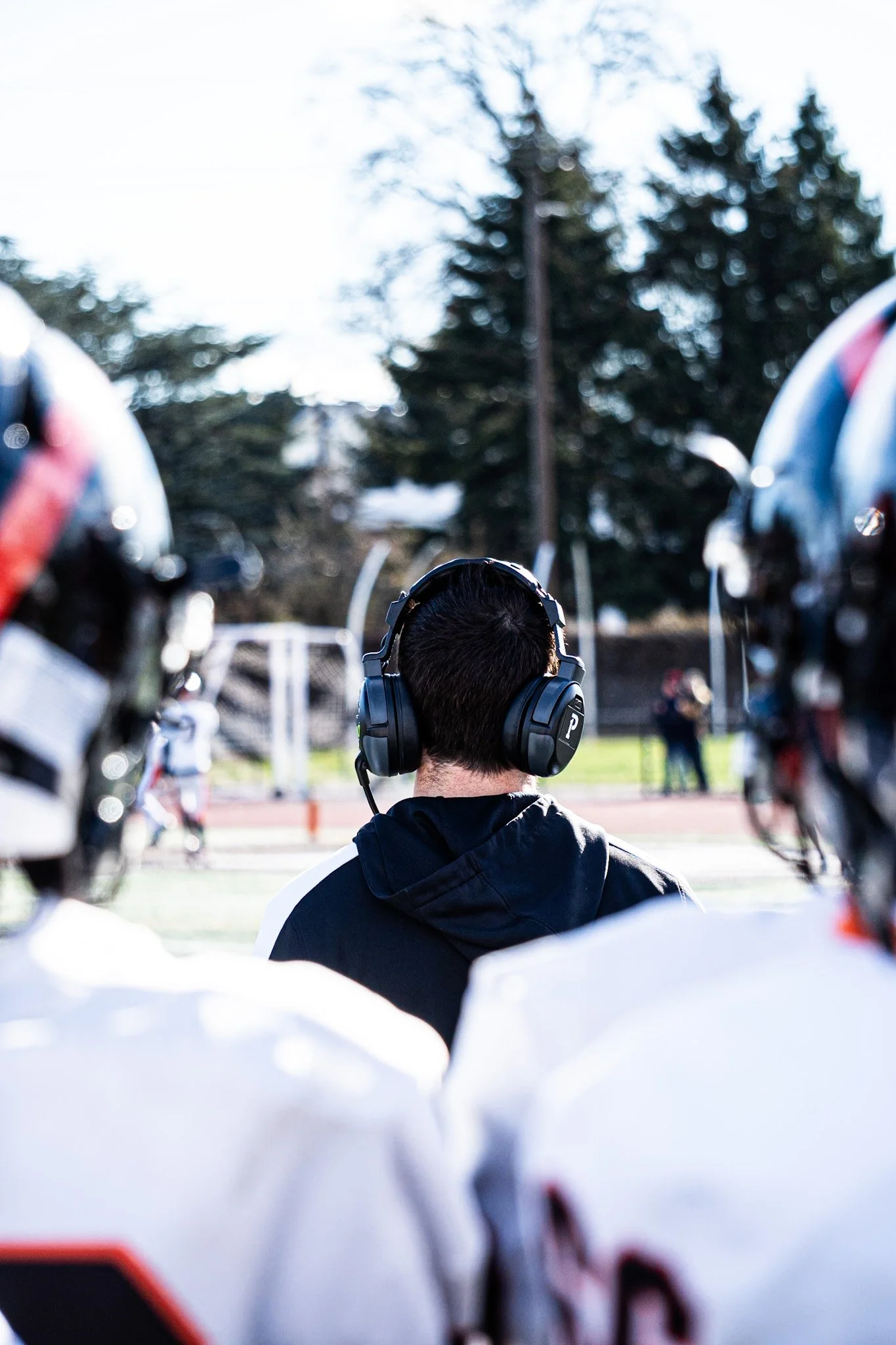 A person with black hair wearing headphones and a black jacket stands outdoors in front of a football field, with trees and a lamppost in the background. The photo is taken from behind and is surrounded by blurred helmets and shoulders of other playe