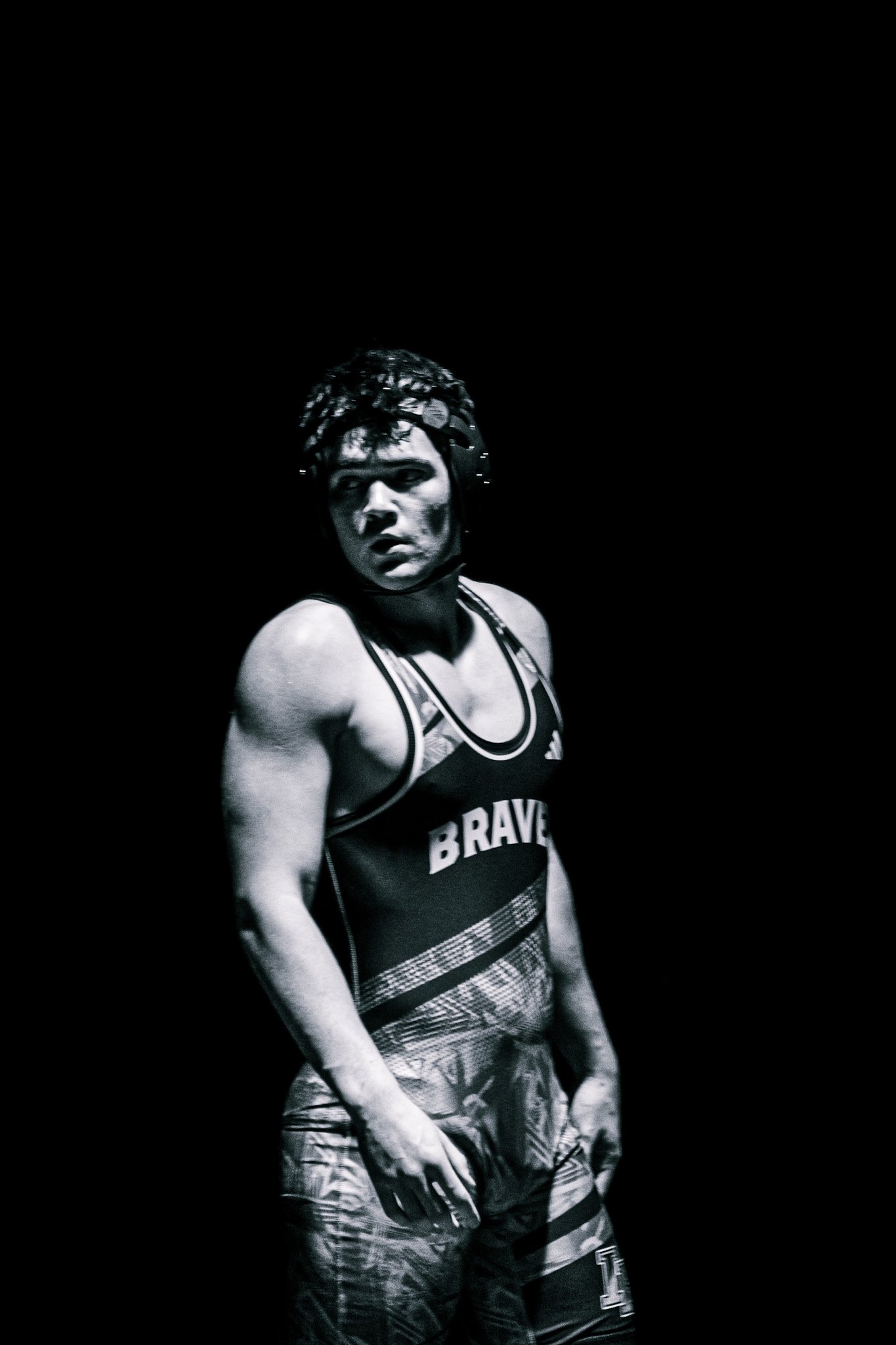 Black and white photo of a female athlete with short curly hair, wearing a sports bra with 'BRAVE' printed on it and patterned shorts, standing against a dark background.