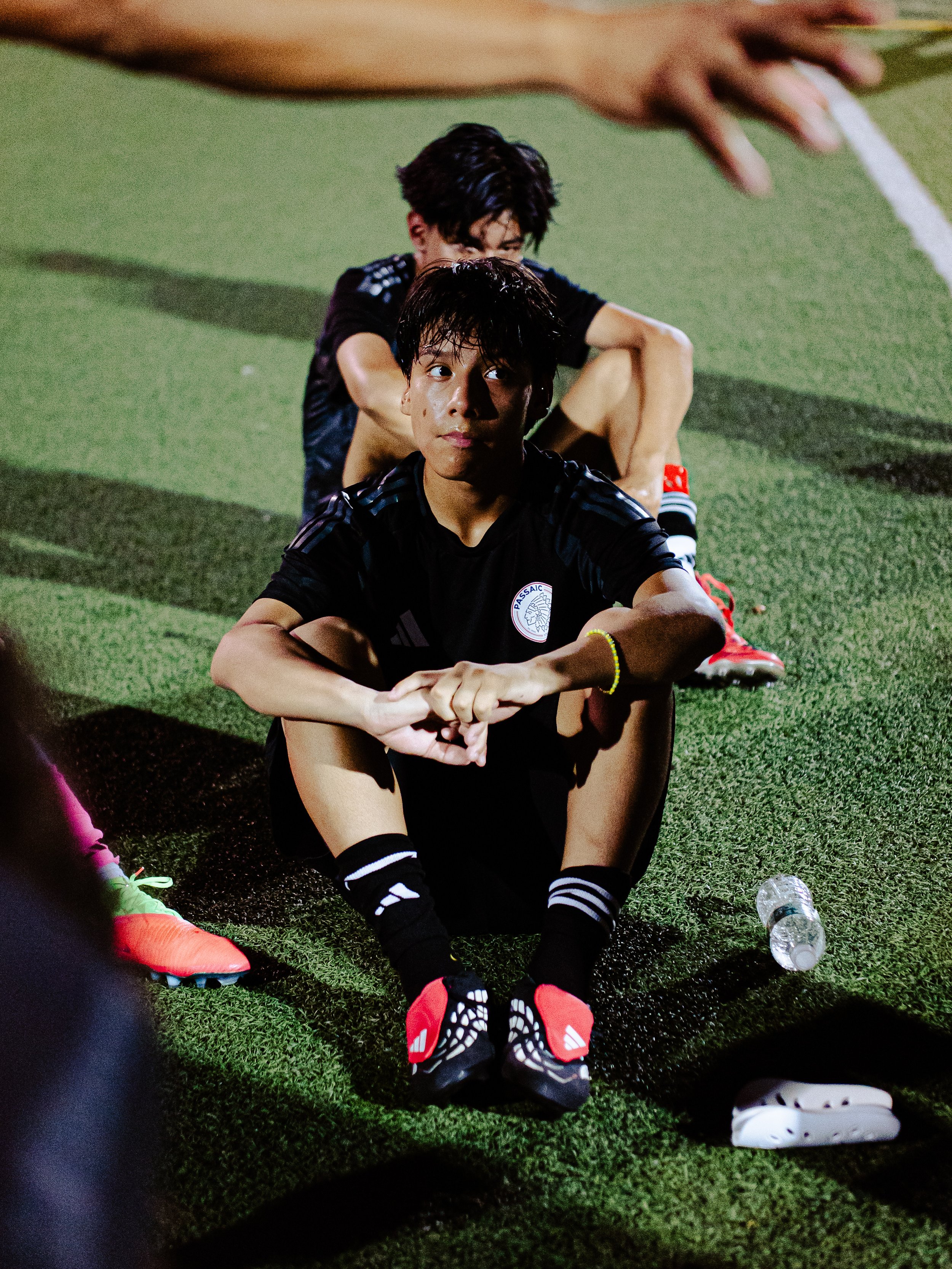 Two young soccer players sitting on a sports field, one in the foreground and one in the background, with water bottles and athletic gear nearby.