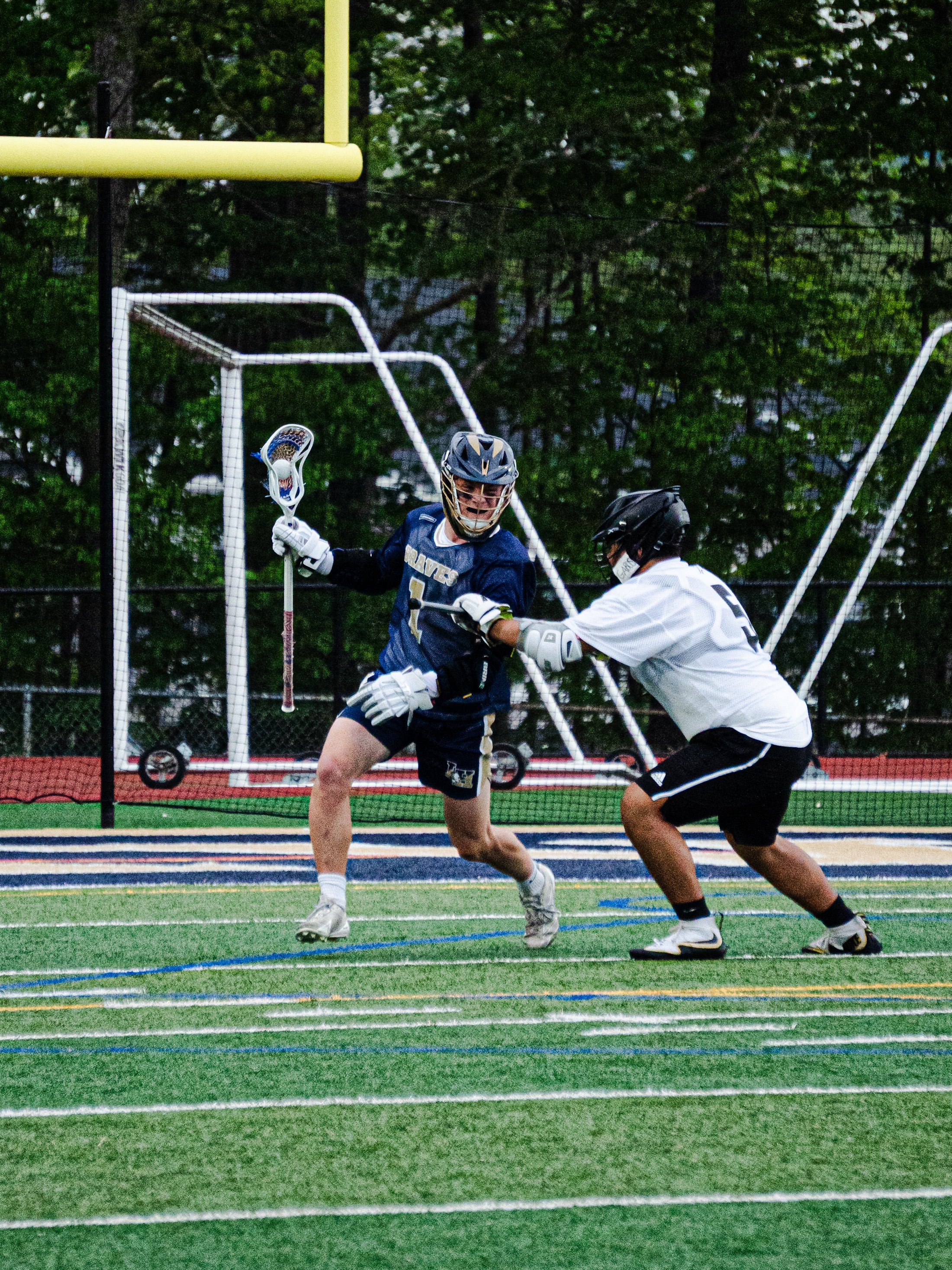 Two lacrosse players on a field during a game, one in a blue uniform with a helmet holding a lacrosse stick, and the other in a white shirt and black shorts. Green trees are in the background.
