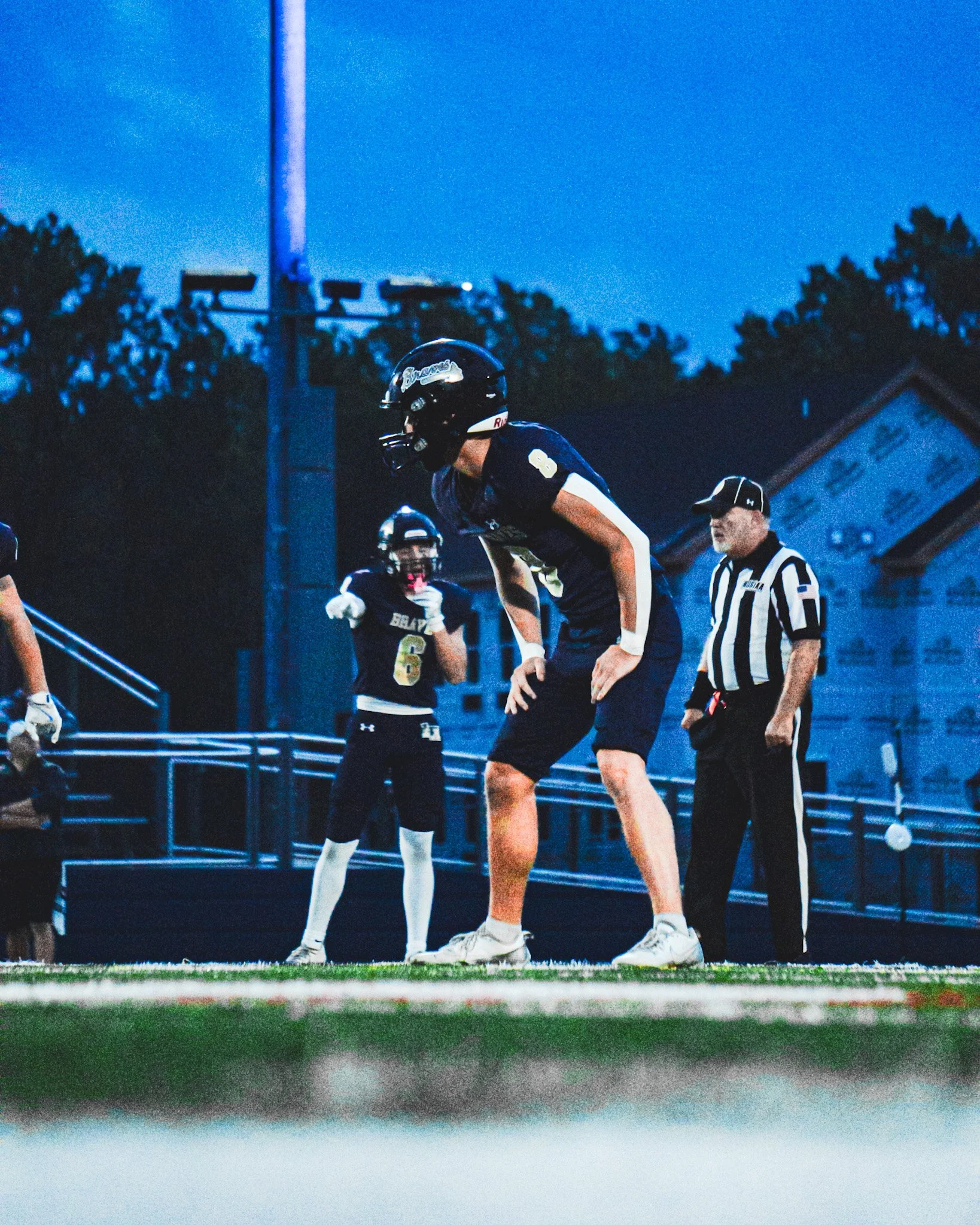 American football players wearing dark jerseys with numbers 8 and 6, standing on the field with referees, under a darkening evening sky.