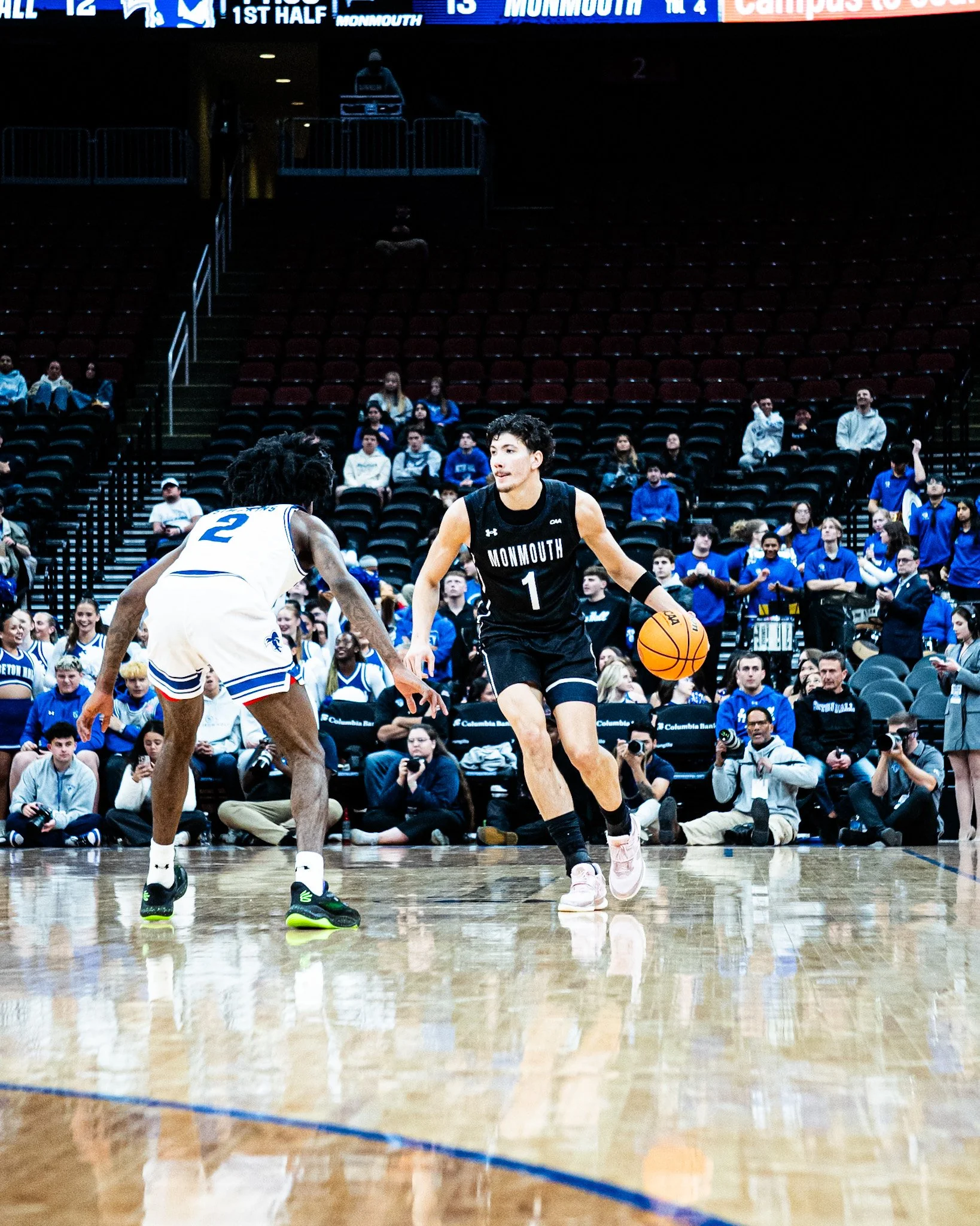 Two basketball players are on the court during a game, one wearing a white jersey with the number 2 and the other wearing a black jersey with the number 1. Spectators watch from the stands, and a scoreboard shows the game is in the first half.