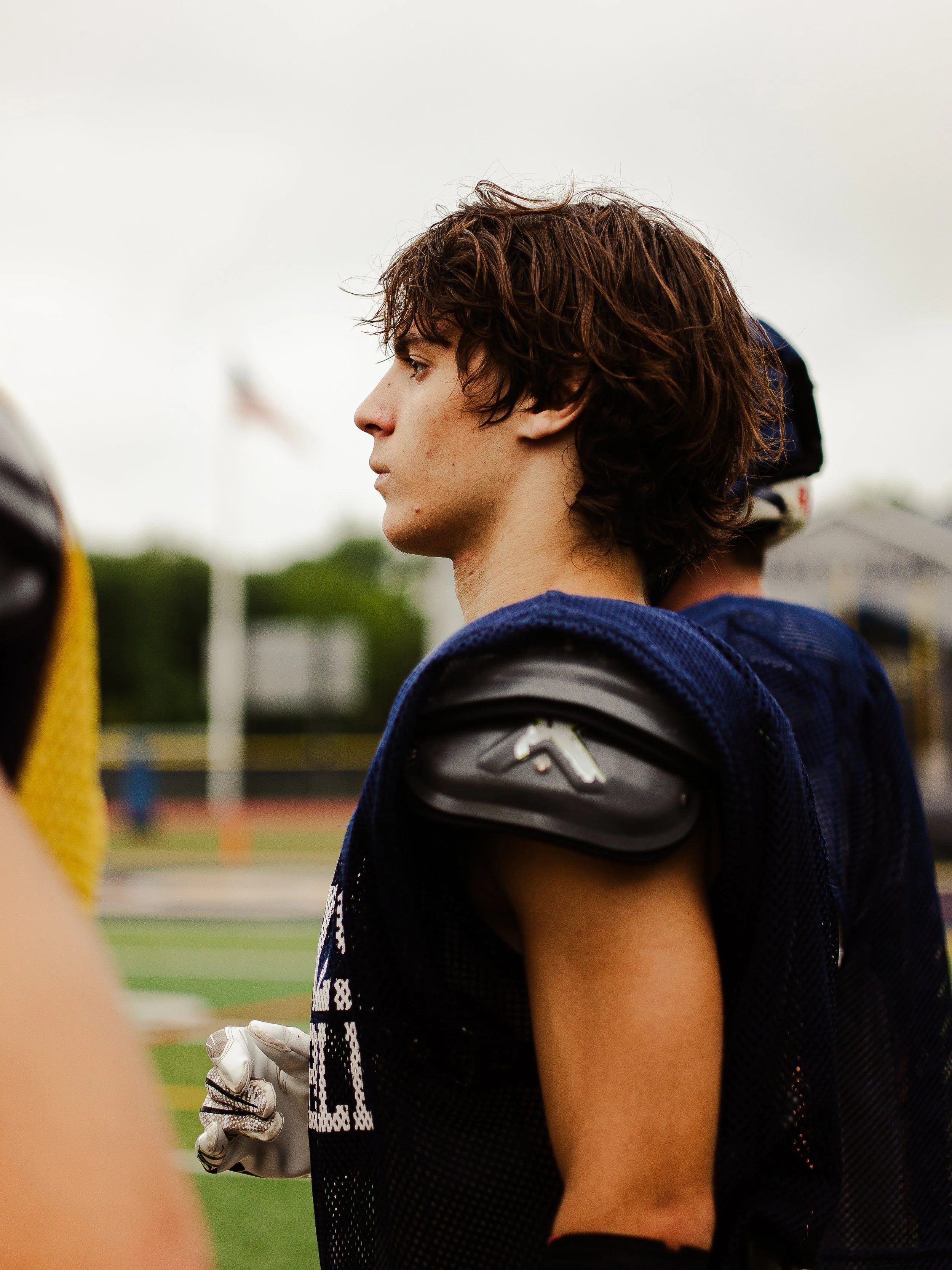 A young male football player with brown curly hair stands on a football field, wearing a blue and black jersey, a football helmet held in his right arm, and a protective shoulder pad. The background shows a blurred sports field, and a person in a yel