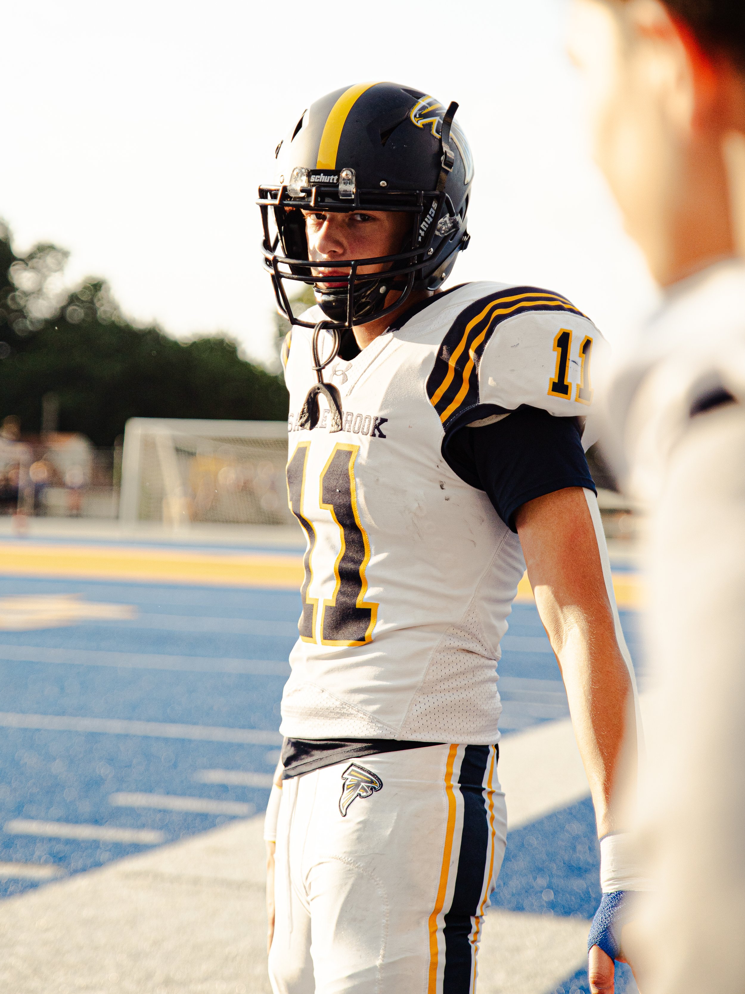 A football player in a black and yellow helmet and white jersey with yellow and black stripes, number 11, standing on a football field during sunset.