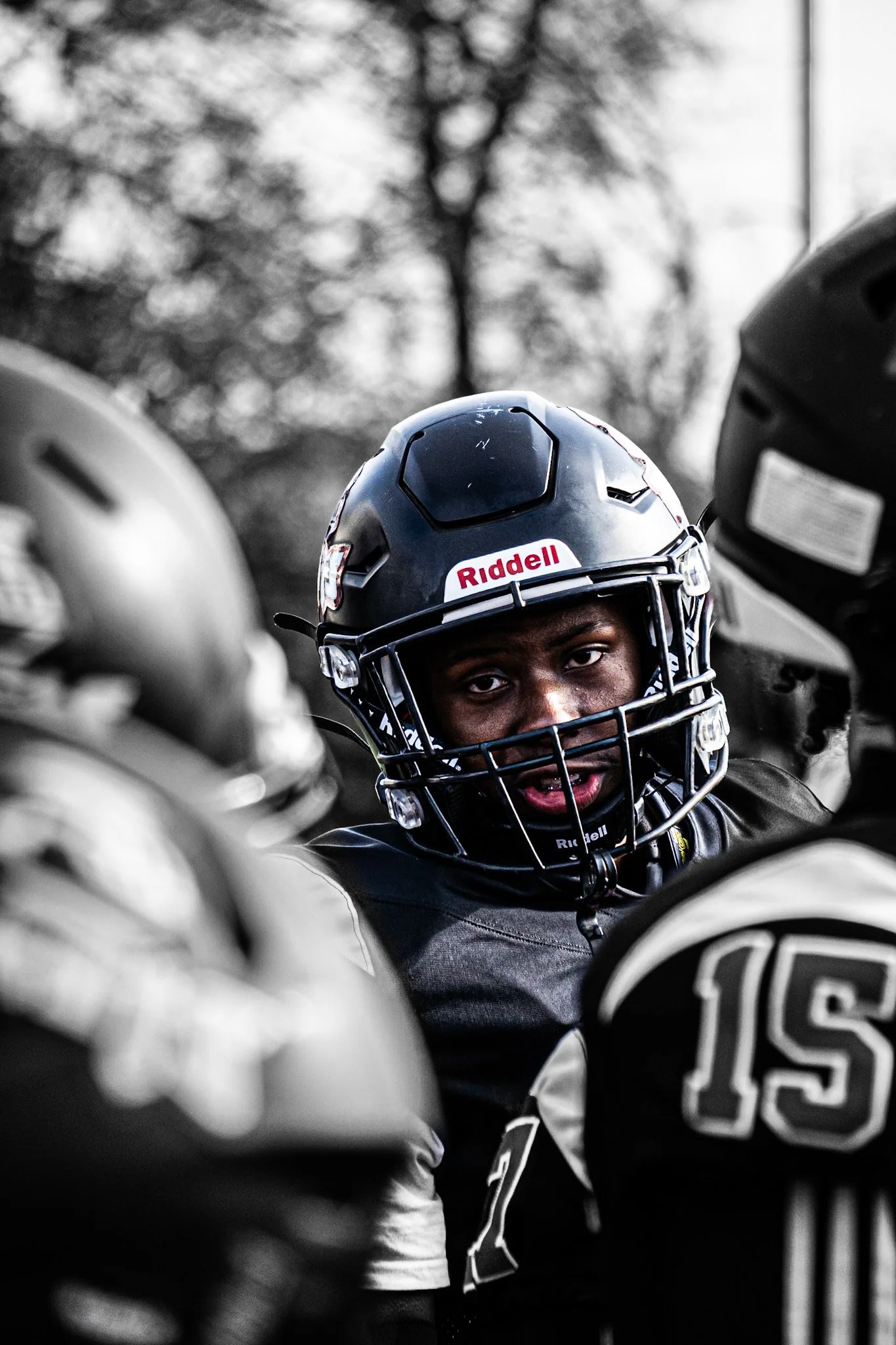 A person wearing a dark football helmet with a faceguard, with a partially visible expression, surrounded by other players in helmets, during a game or practice outdoors.