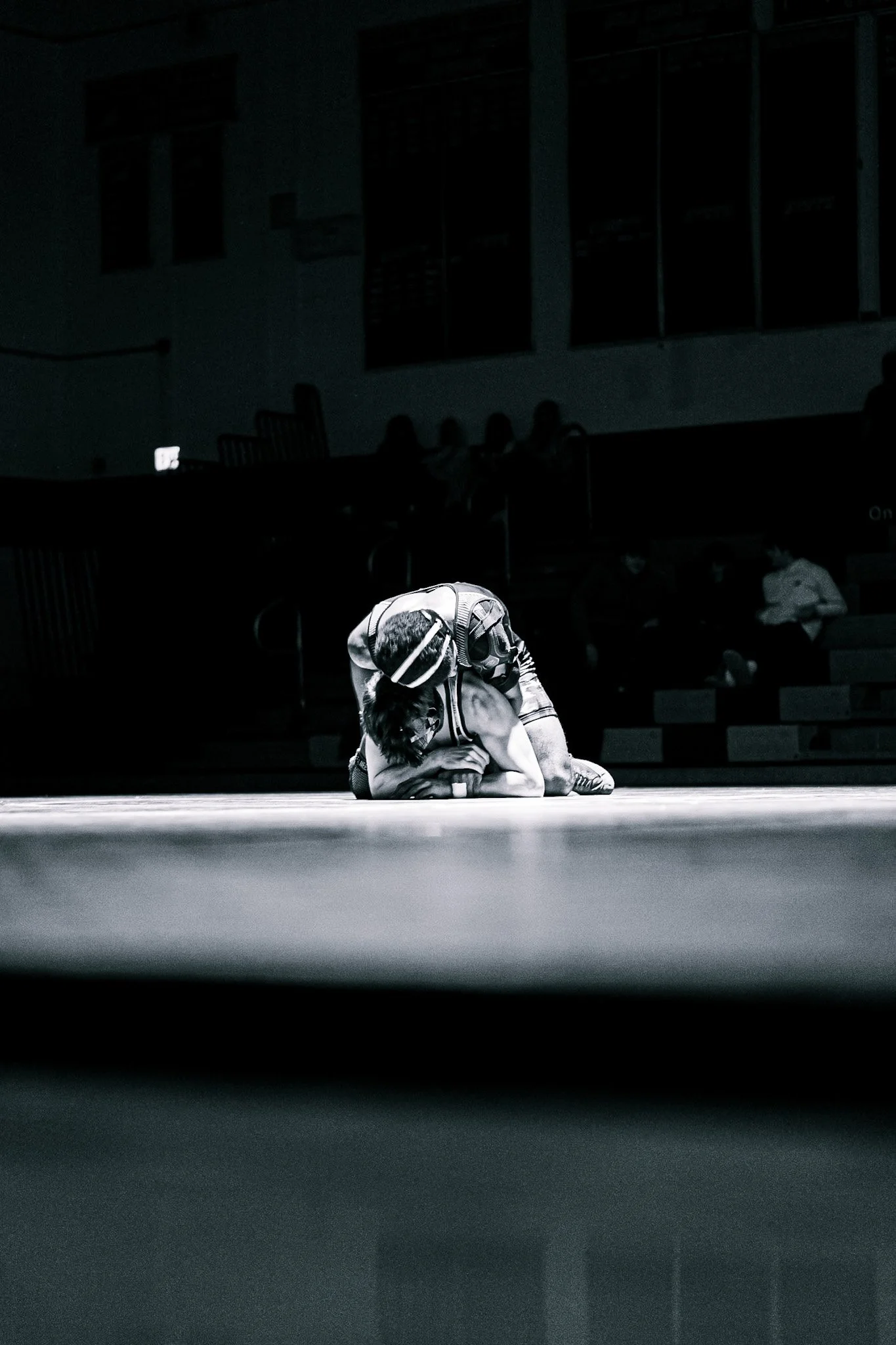 A young person sitting on a gymnasium floor, resting with their head bowed and hands covering their face, wearing a backpack and athletic clothing, with a dark background and faint shadowy figures in the distance.