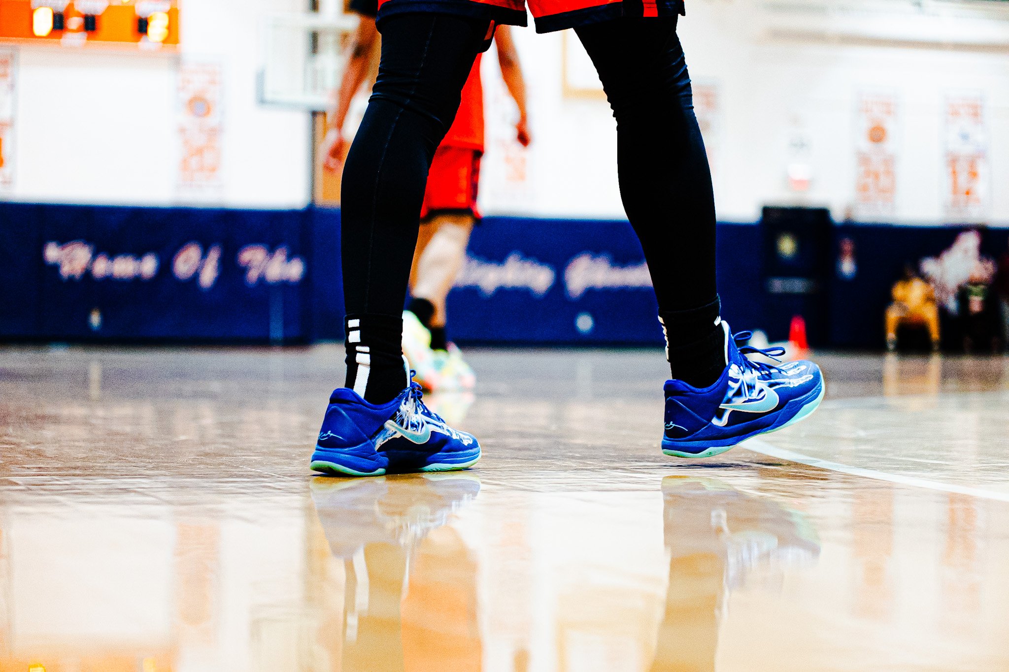 Close-up of a person wearing blue athletic shoes standing on a polished gymnasium floor, with another player blurred in the background.