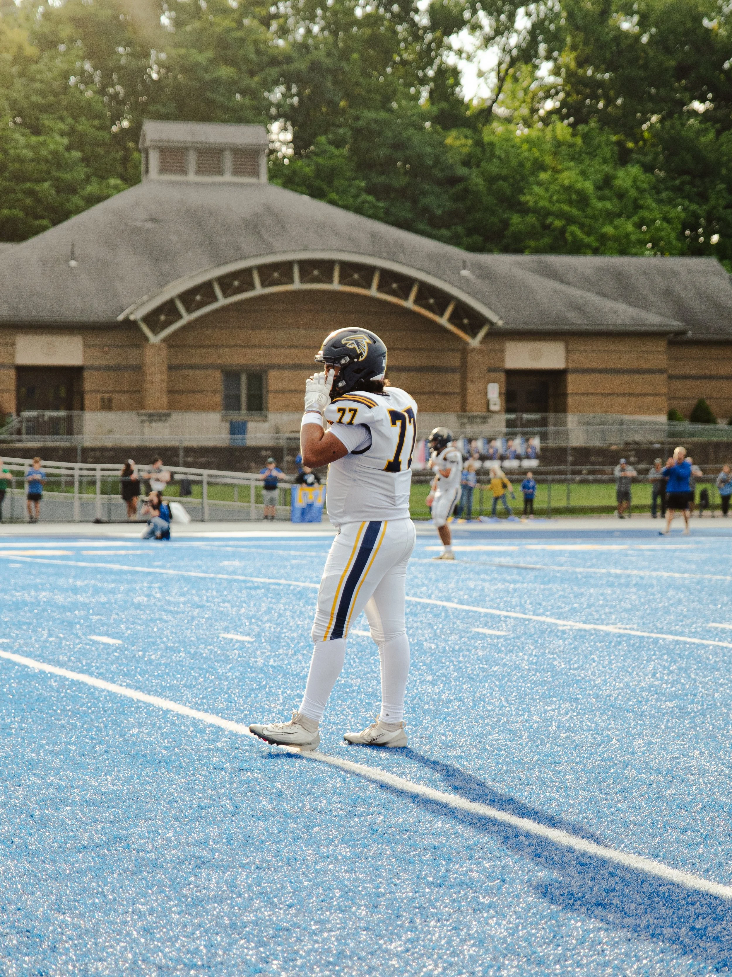 An American football player wears a white jersey with yellow and black accents, standing on a blue-colored football field, holding a helmet and talking on a mobile phone, with a school building and trees in the background.