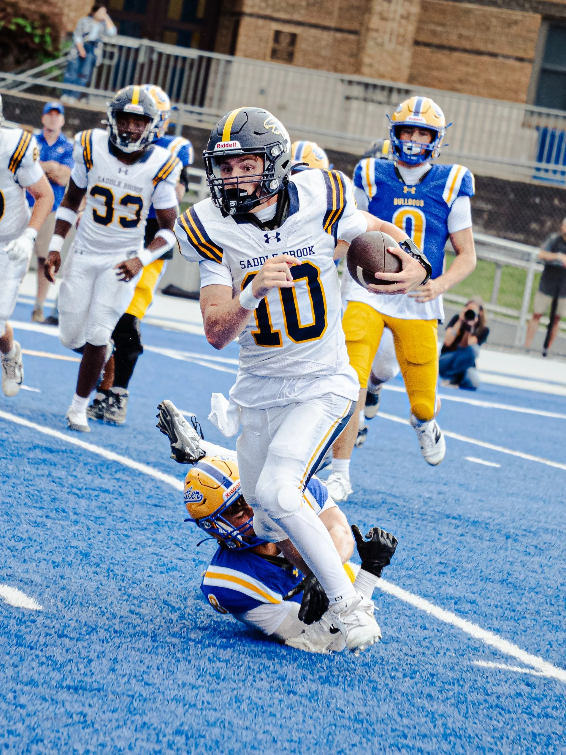 A football player in a white and yellow uniform runs with the ball, tackled by a player in a blue and yellow uniform, on a blue field during a game.