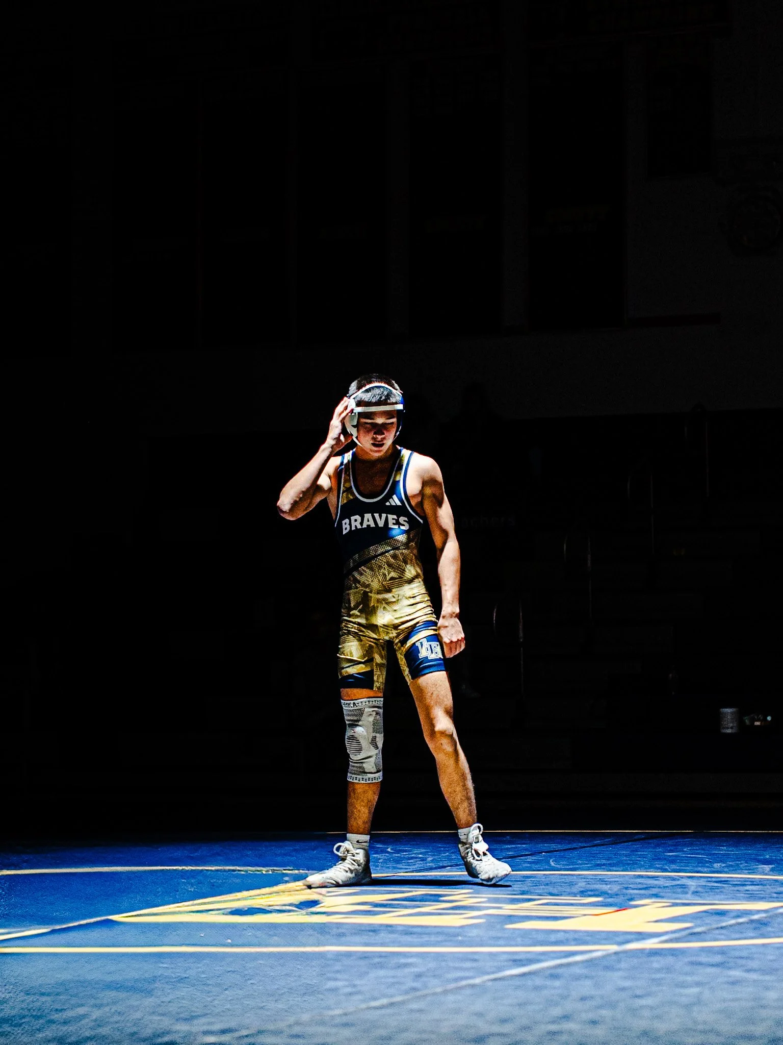 A young male wrestler wearing a singlet and a protective headgear, standing on a wrestling mat in a gymnasium with dark background lights.