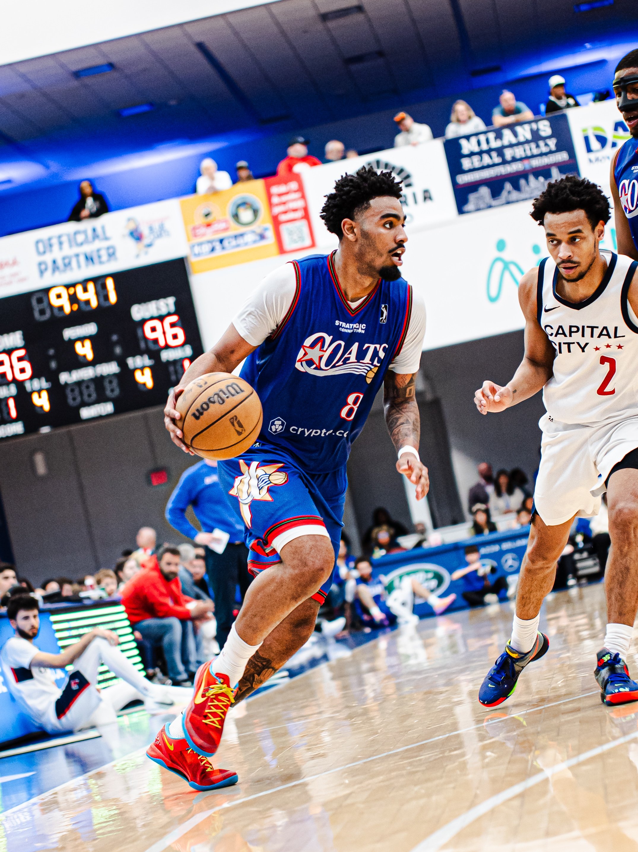 Two basketball players in action during a game. The player in a blue jersey with red and yellow sneakers has possession of the ball, while the player in a white jersey with black and red sneakers defends. Spectators are in the background.