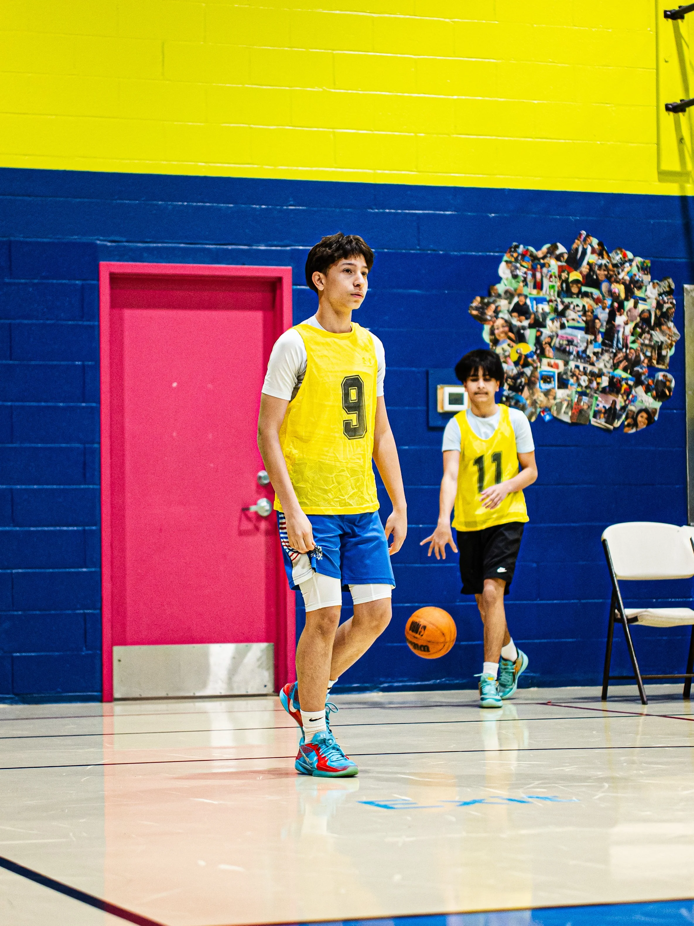 Young male basketball player in a yellow jersey with the number 9 dribbling an orange basketball on an indoor court with blue and yellow walls. Another player in a yellow jersey with the number 11 is in the background.