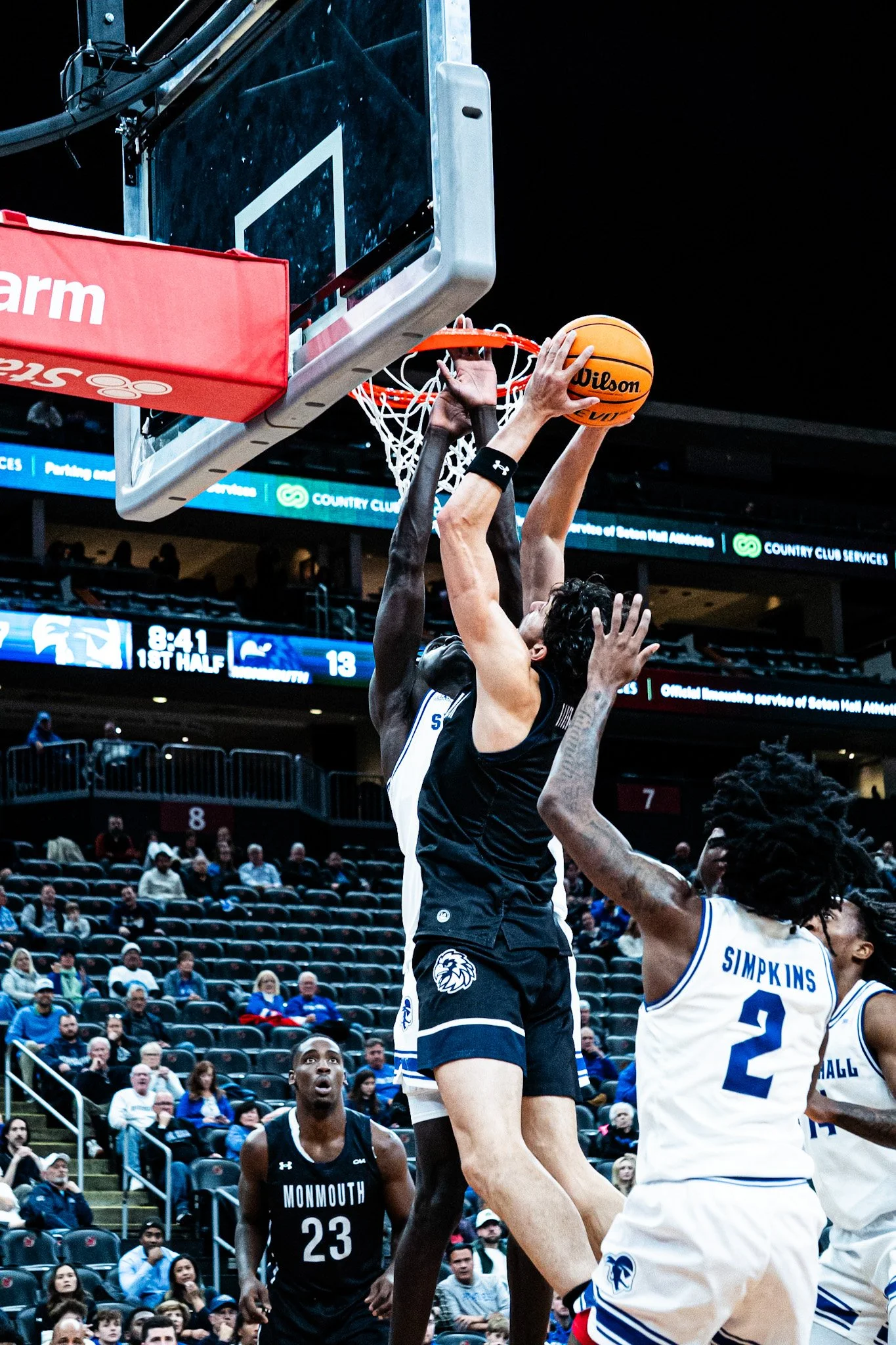 Basketball player in black uniform jumps to make a shot as a player in white attempts to block the shot during a game, with spectators in the background.